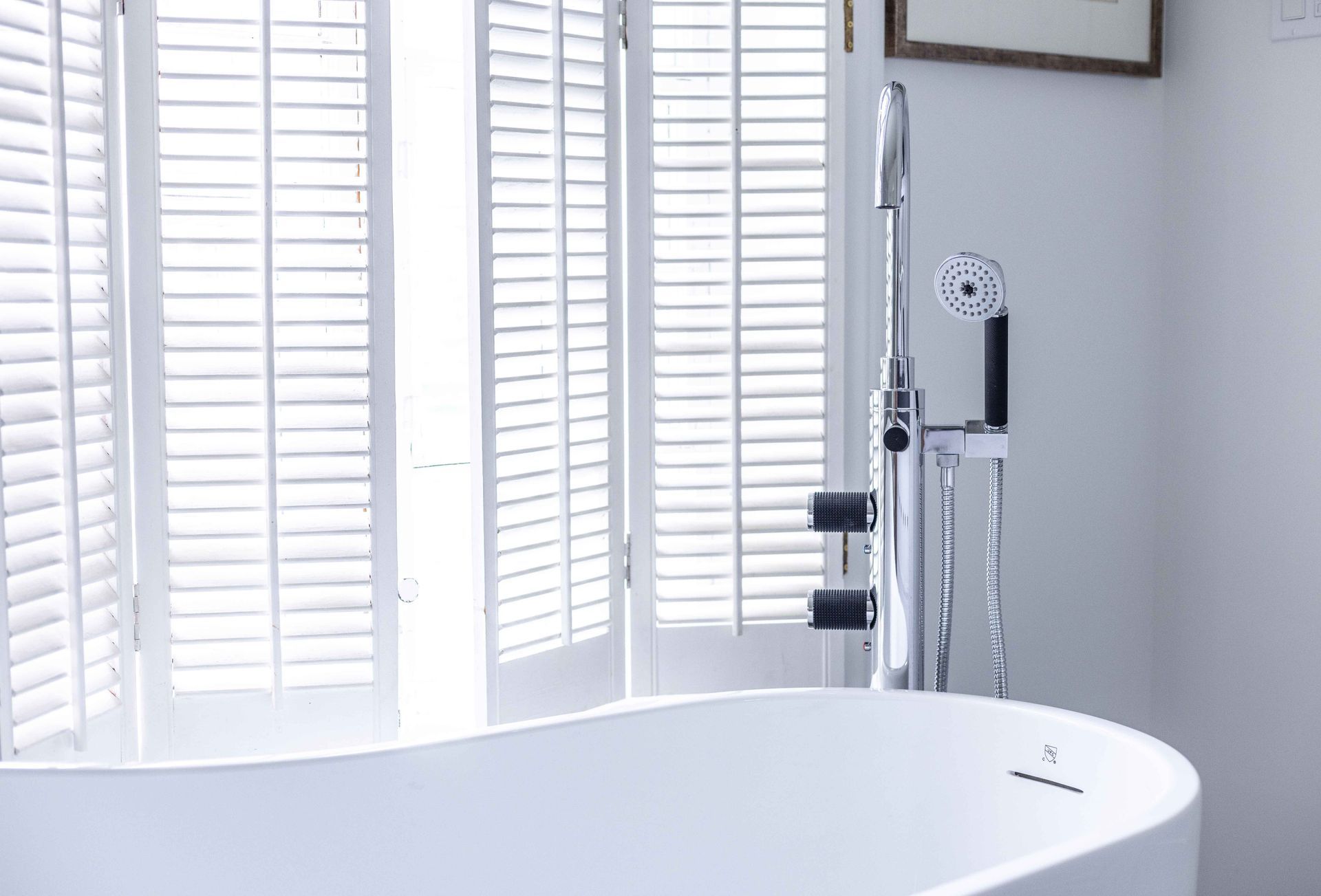 Baignoire blanche près de la fenêtre avec volets blancs, robinetterie de douche chromée.