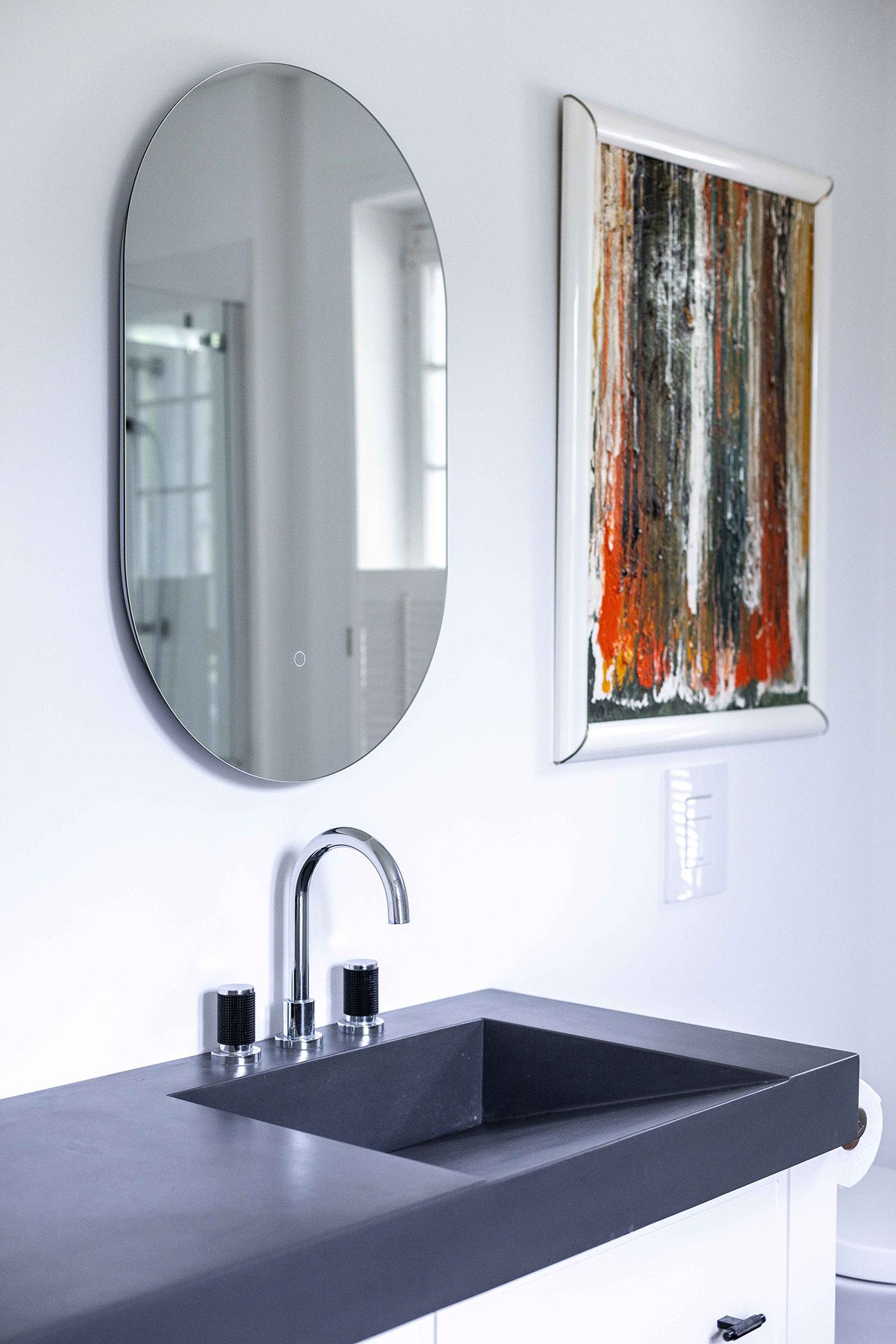 Miroir ovale au-dessus d'un lavabo gris foncé avec robinet argenté dans une salle de bains moderne. Œuvre d'art à droite.