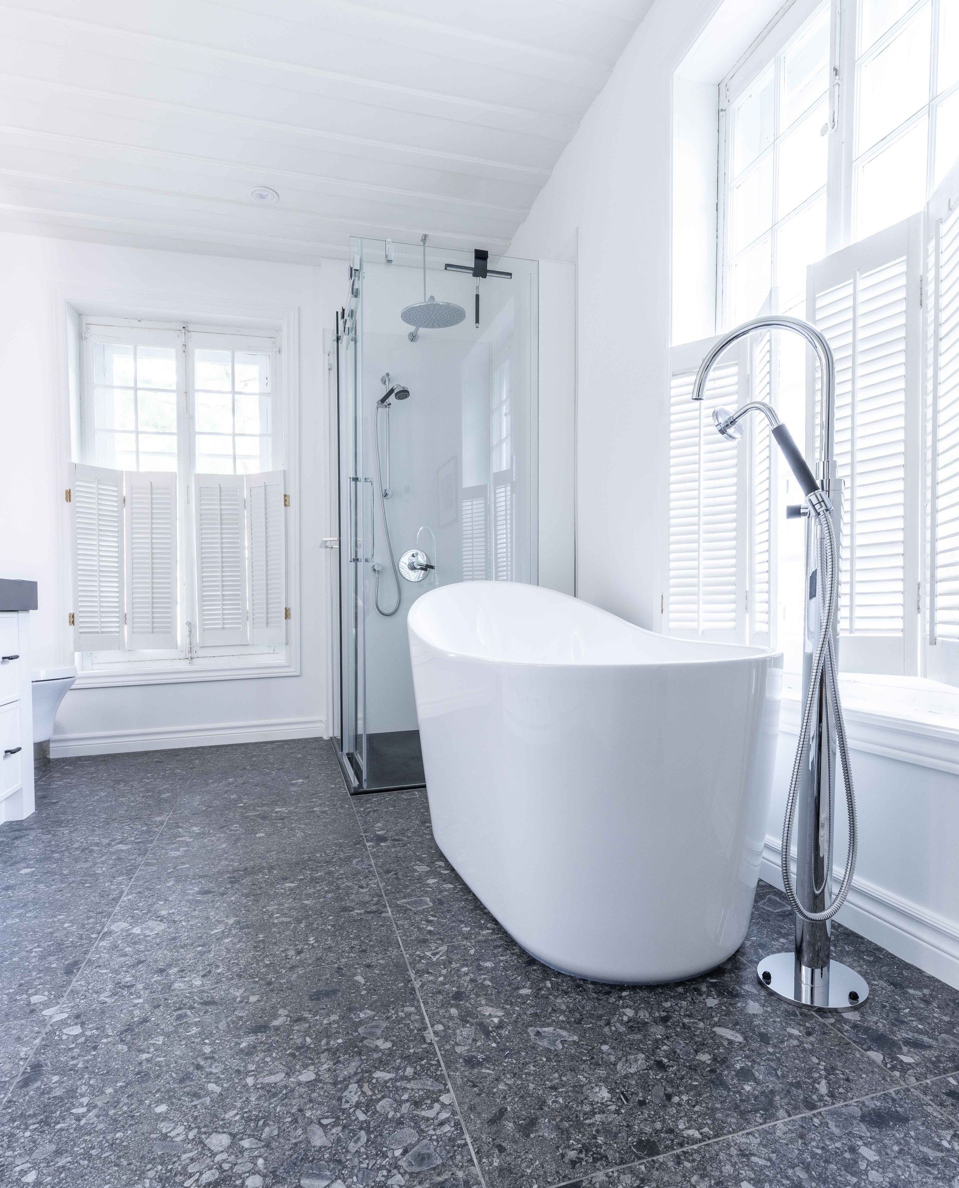 Salle de bains moderne blanche avec baignoire îlot, douche en verre et fenêtres à volets. Sol moucheté noir et gris.