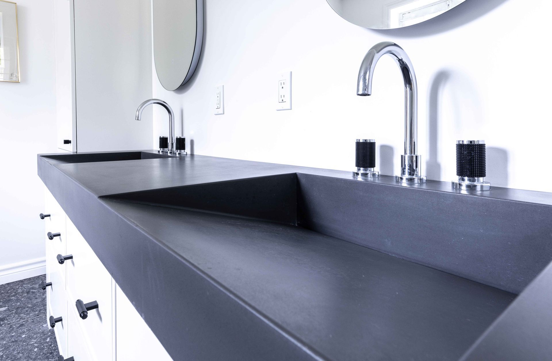 Salle de bains moderne avec comptoir gris foncé, deux lavabos avec robinetterie chromée et armoires blanches.