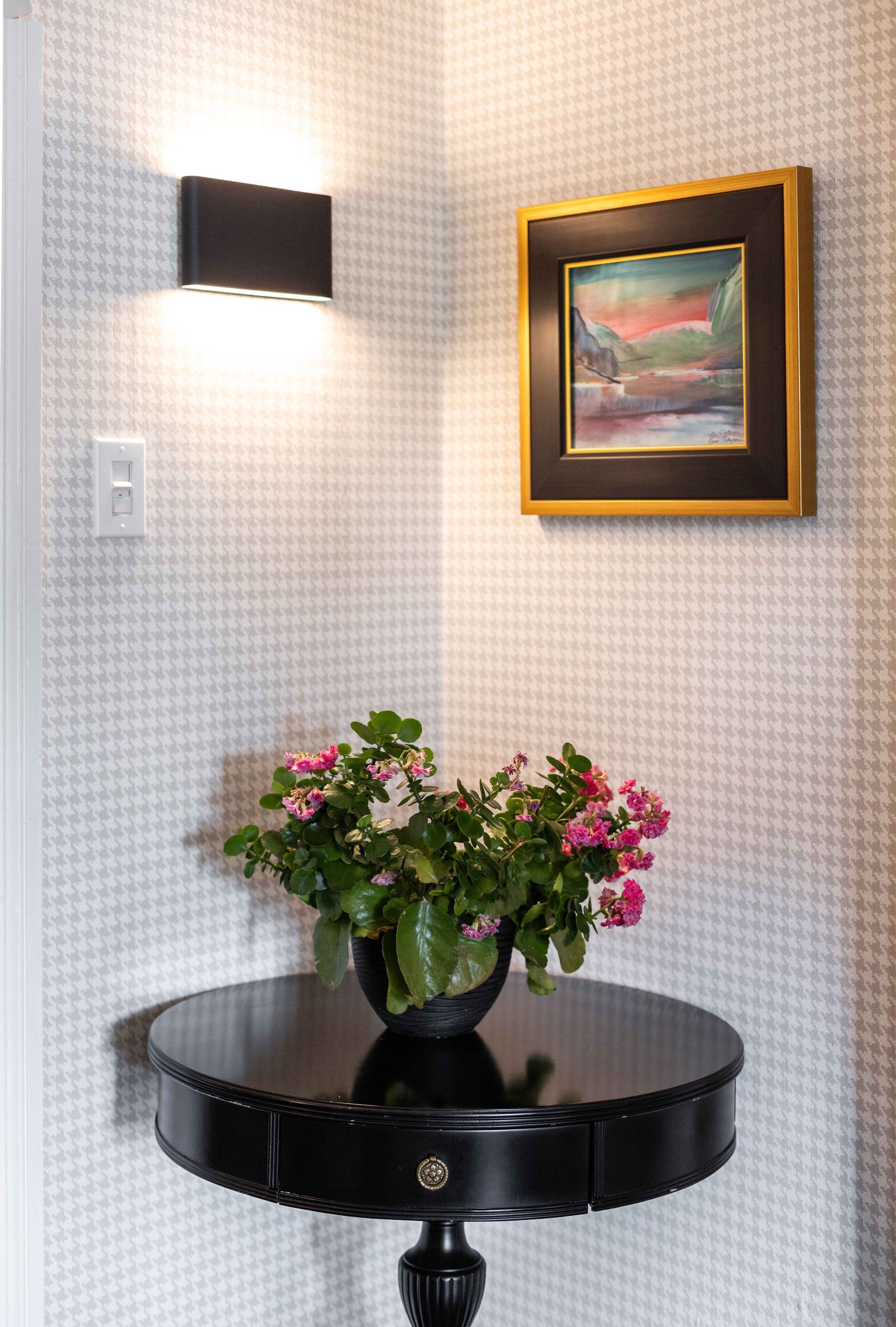 Table noire avec des fleurs dans un coin, sous un paysage encadré et une applique murale.