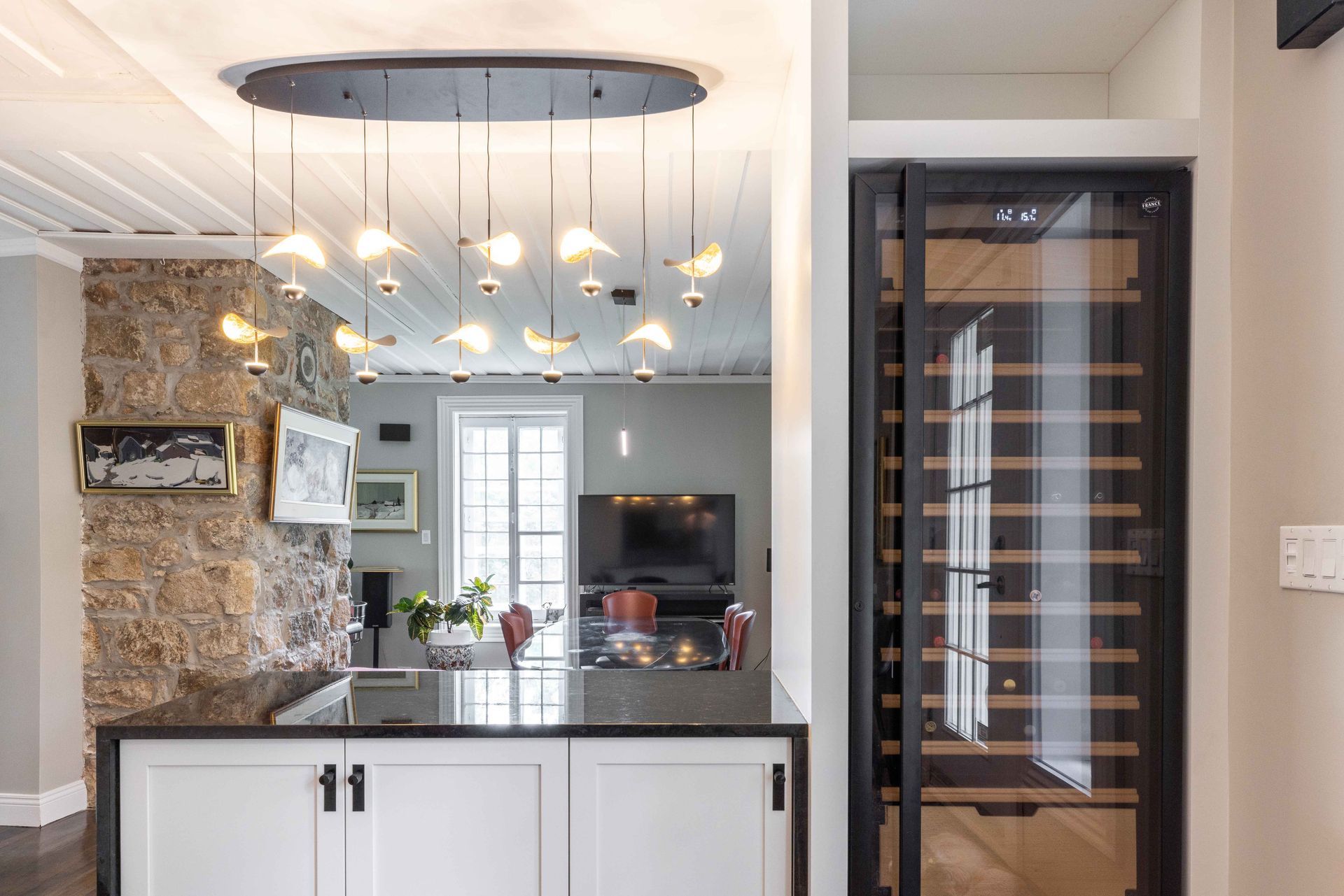 Vue intérieure d'une cuisine moderne avec mur en pierre, lustre, cave à vin et vue sur le salon.