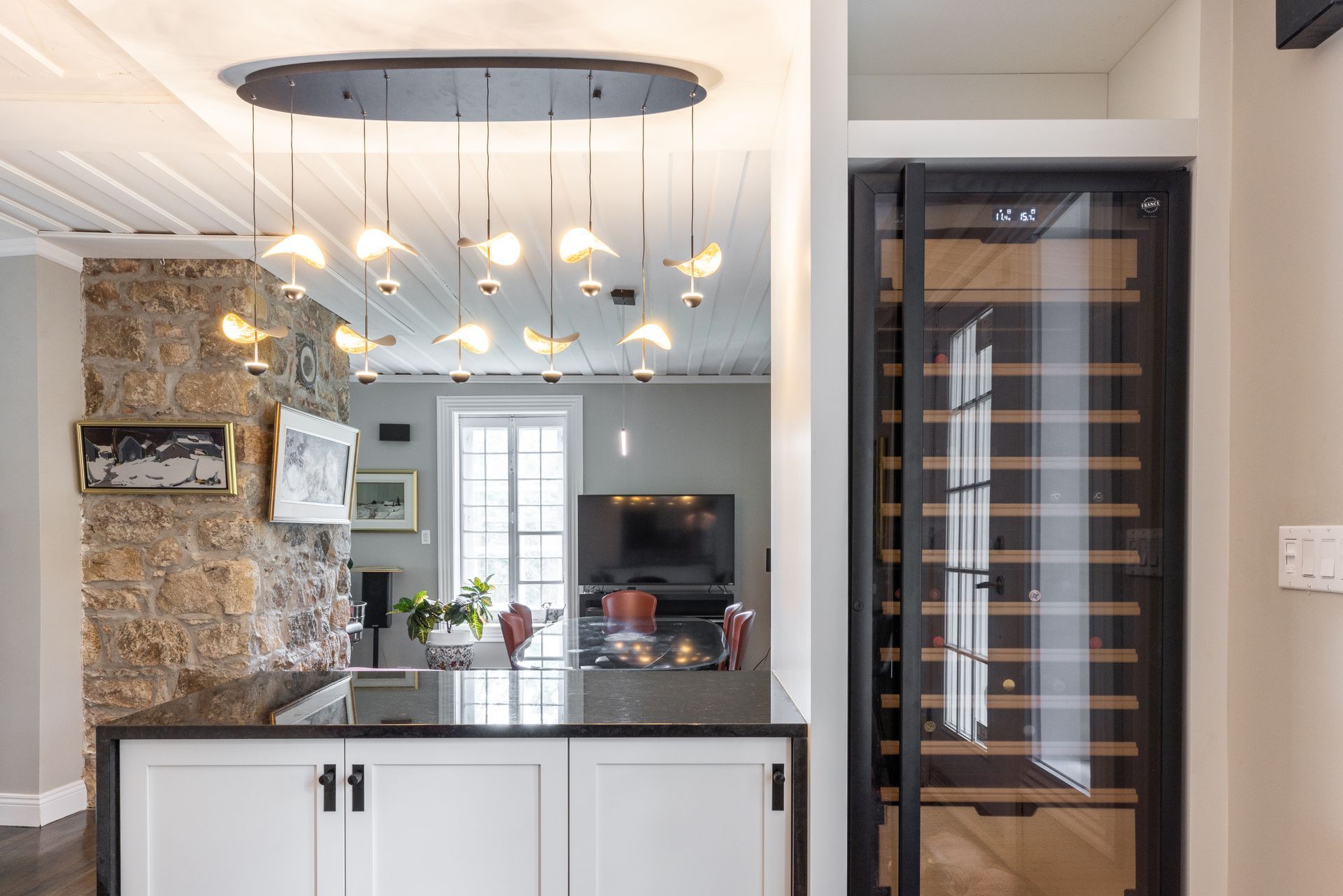 Cuisine avec cave à vin, îlot central avec suspensions lumineuses, mur en pierre et vue sur un salon avec télévision.