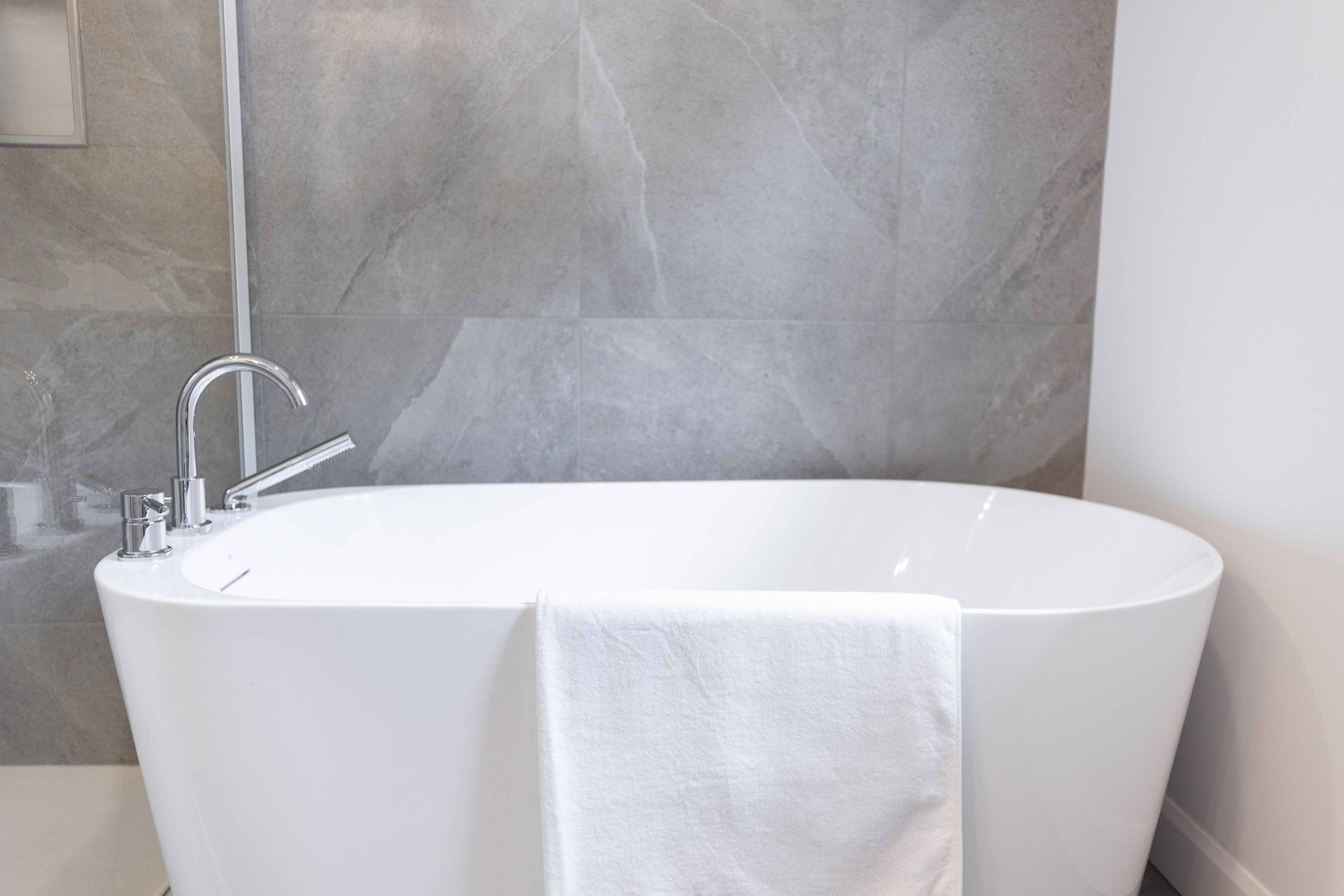 Baignoire ovale avec robinetterie chromée contre un mur carrelé gris. Une serviette blanche est accrochée à la baignoire.