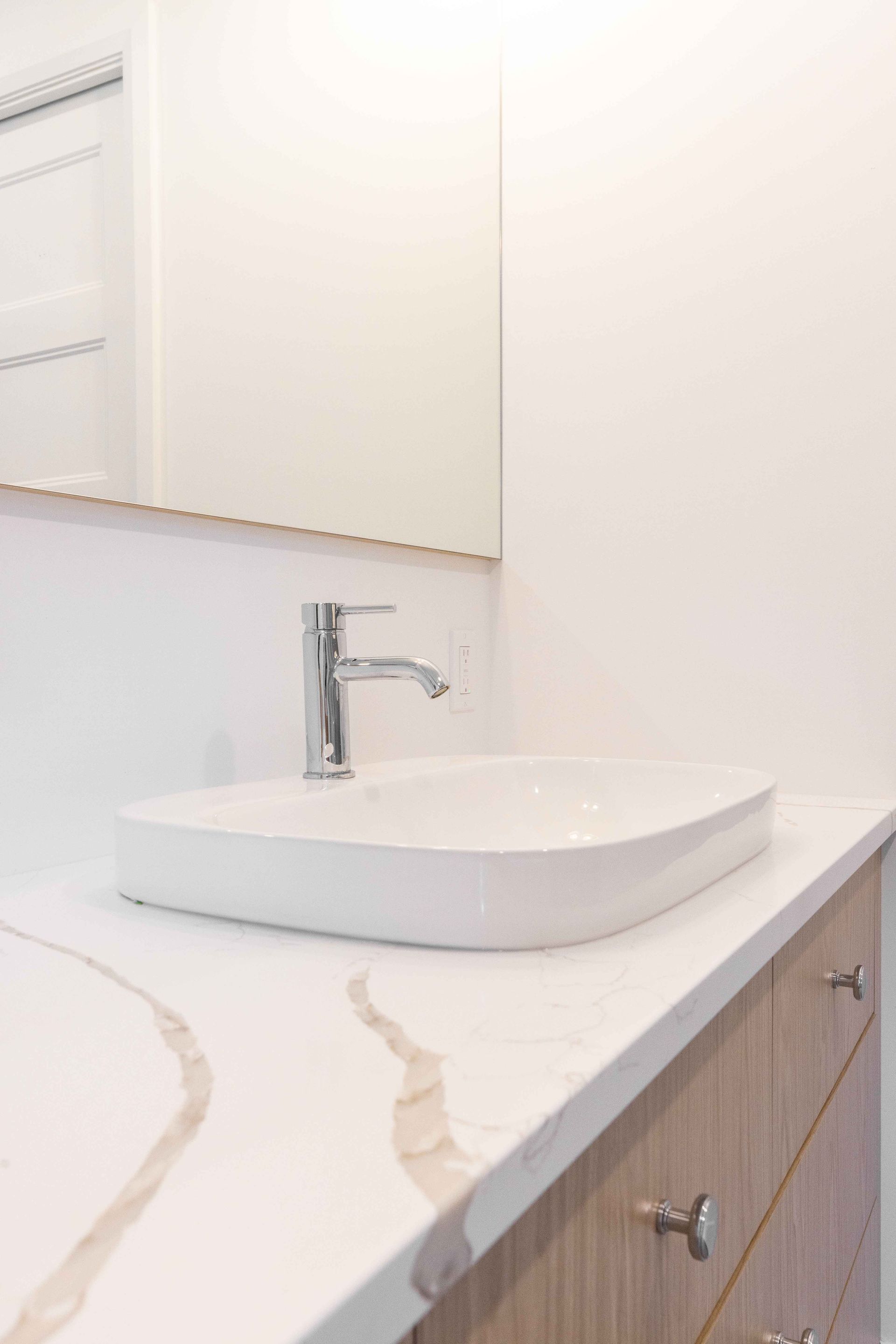 Salle de bains moderne avec lavabo, robinet et plan de travail blancs, meubles en bois et miroir.