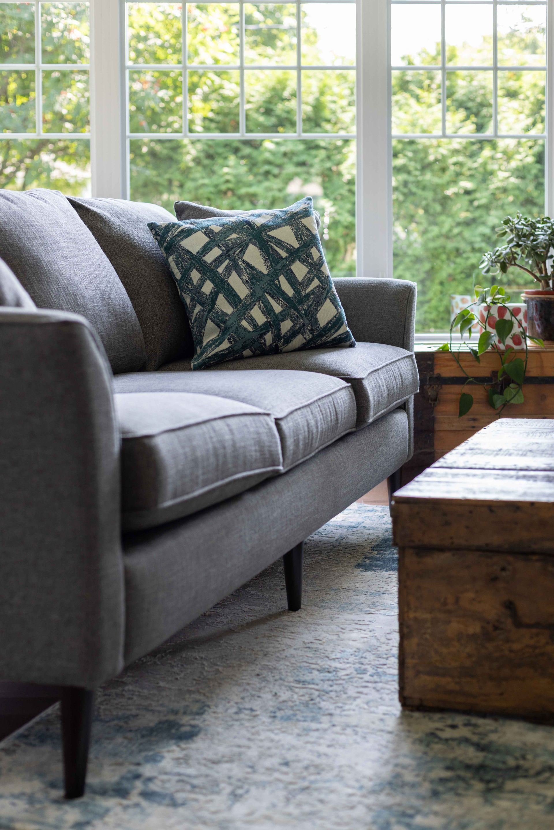 Canapé gris avec coussin à motifs, à côté d'un coffre et d'une fenêtre avec de la verdure.