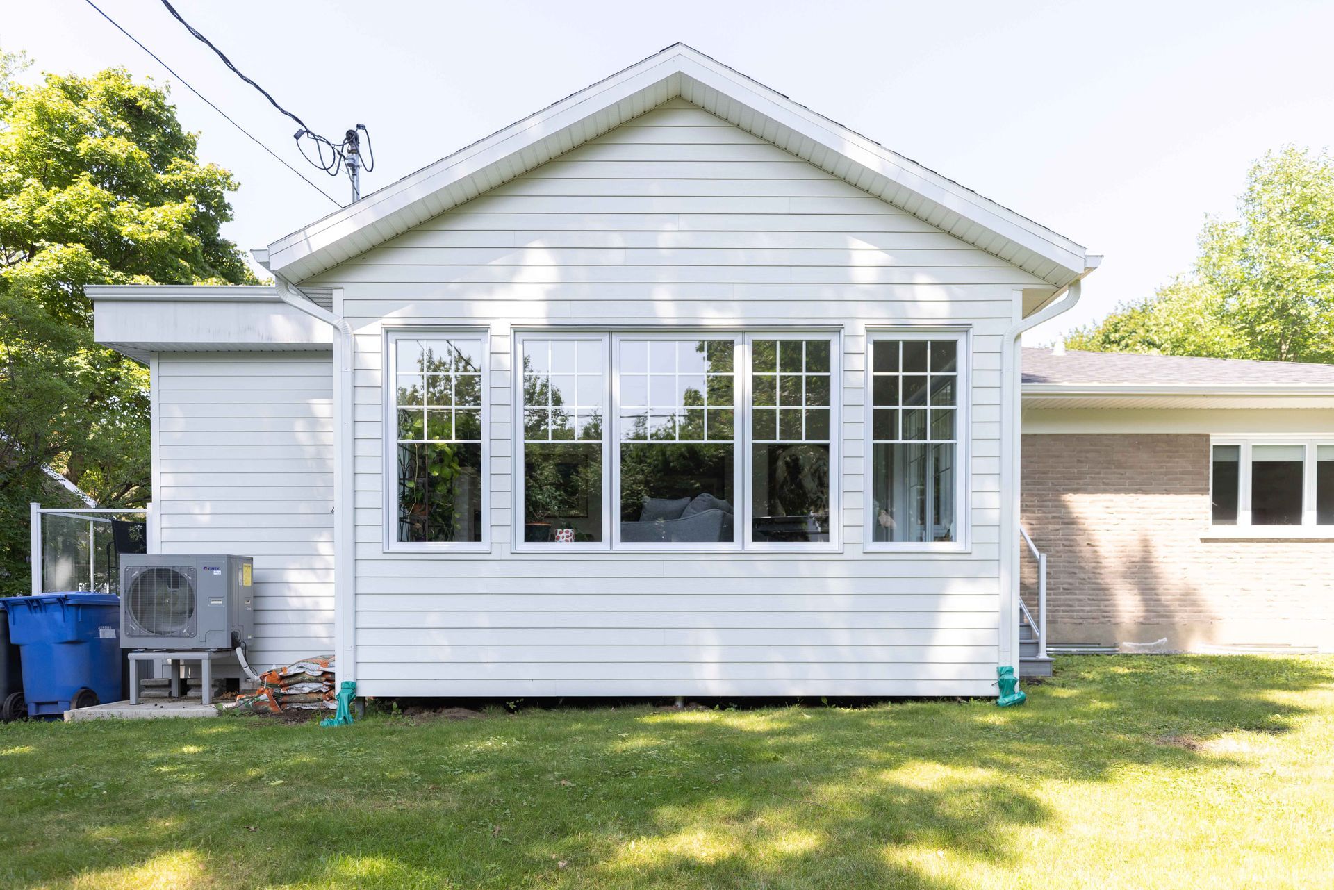 Extension de maison blanche avec de nombreuses fenêtres ; pelouse au premier plan, arbres et ciel visibles.