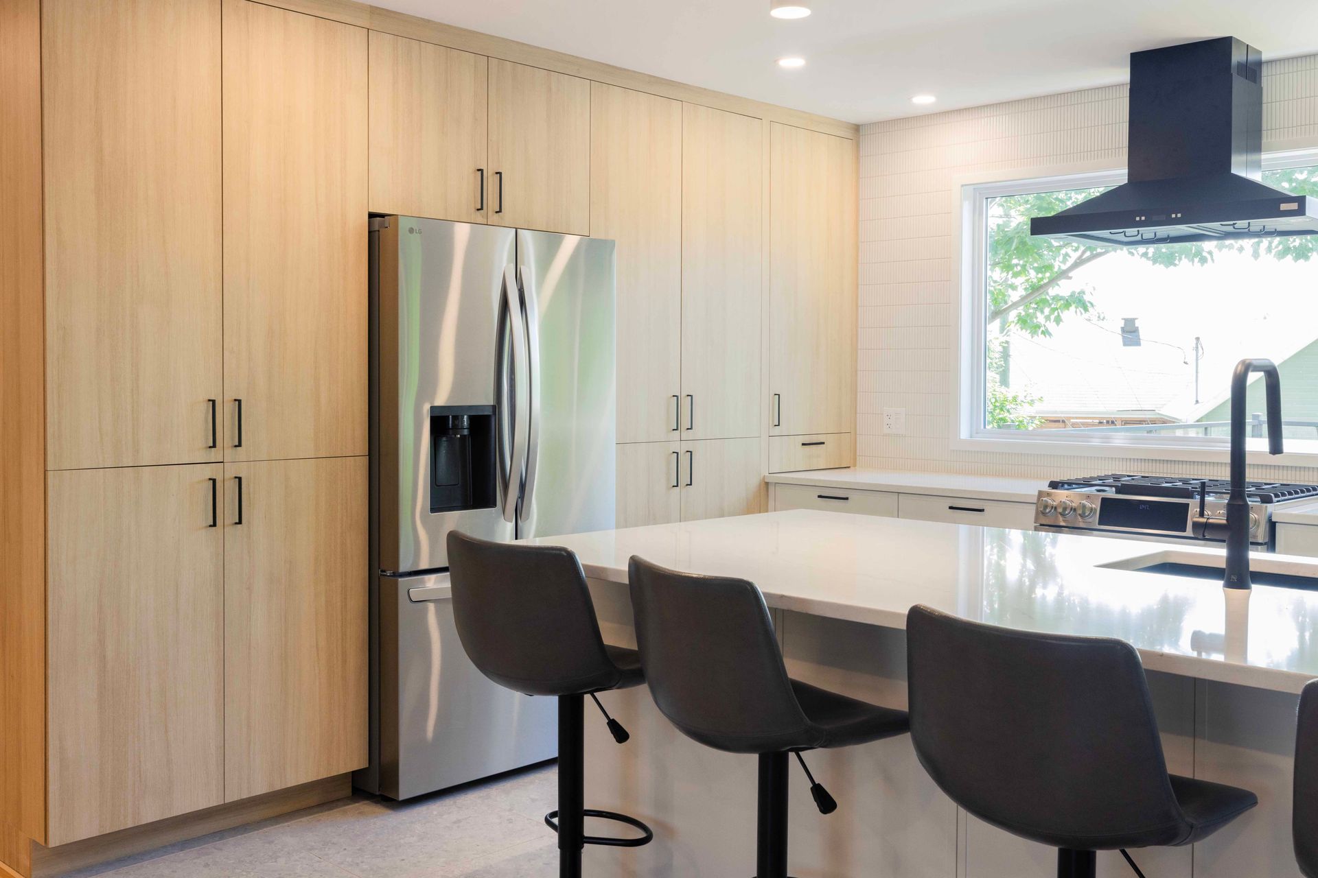 Cuisine moderne avec armoires en bois clair, réfrigérateur en acier inoxydable, îlot central avec tabourets gris et hotte aspirante noire.