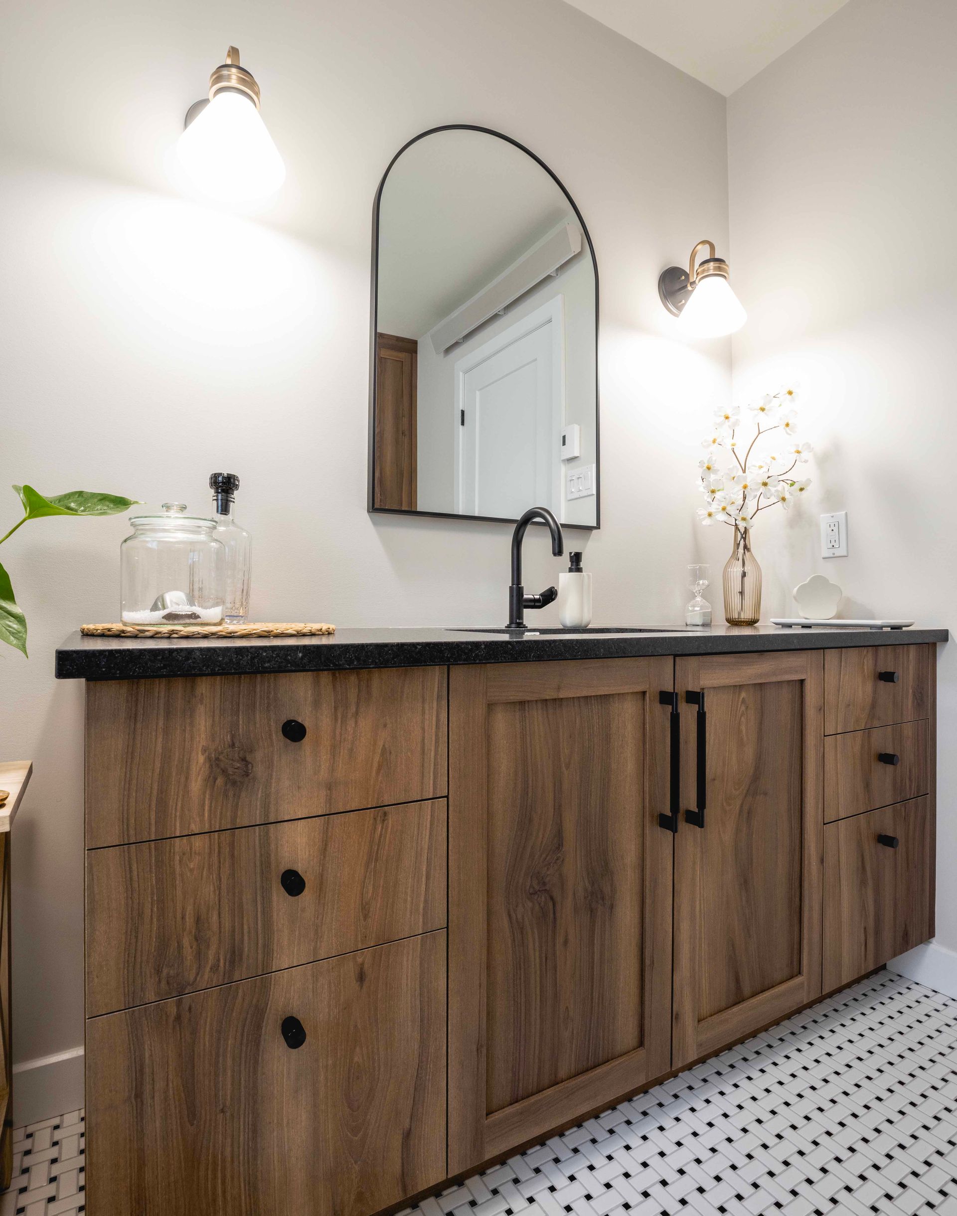 Salle de bains avec meuble-lavabo en bois, comptoir noir, miroir arqué et appliques murales.