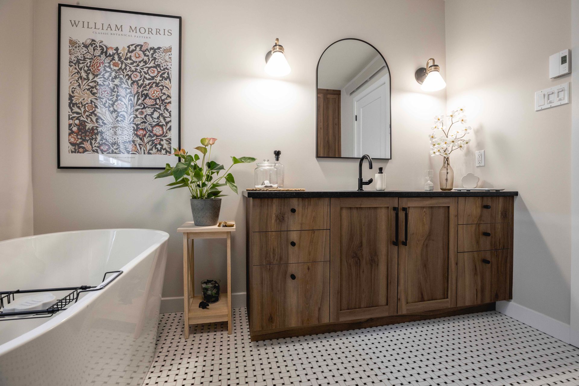 Salle de bains avec meuble-lavabo en bois, miroir arqué, baignoire, reproduction d'art et carrelage noir et blanc au sol.