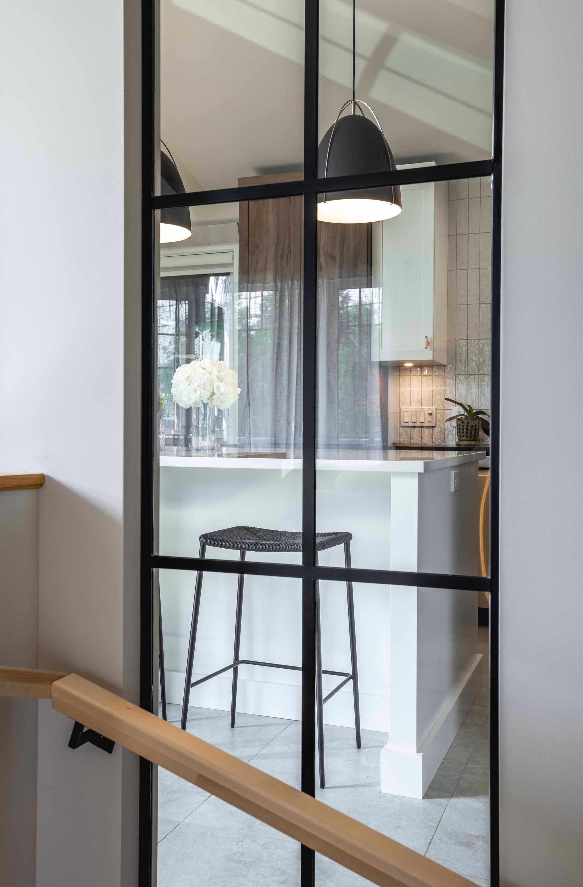 Porte vitrée à cadre noir dévoilant une cuisine avec un tabouret de bar, un plan de travail blanc et une suspension lumineuse.