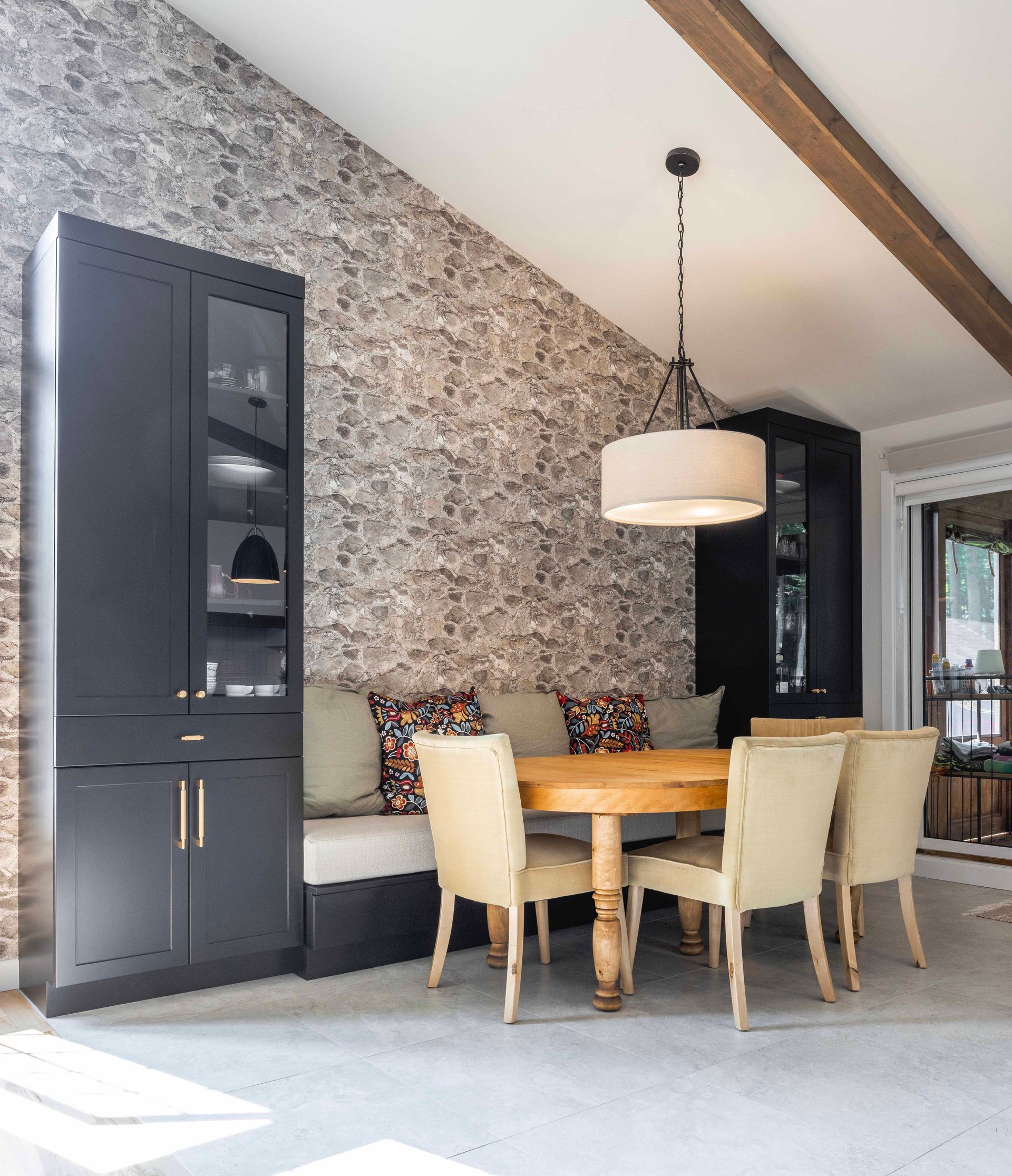 Coin repas avec mur en pierre, meubles intégrés noirs, table ronde, chaises claires et luminaires suspendus.