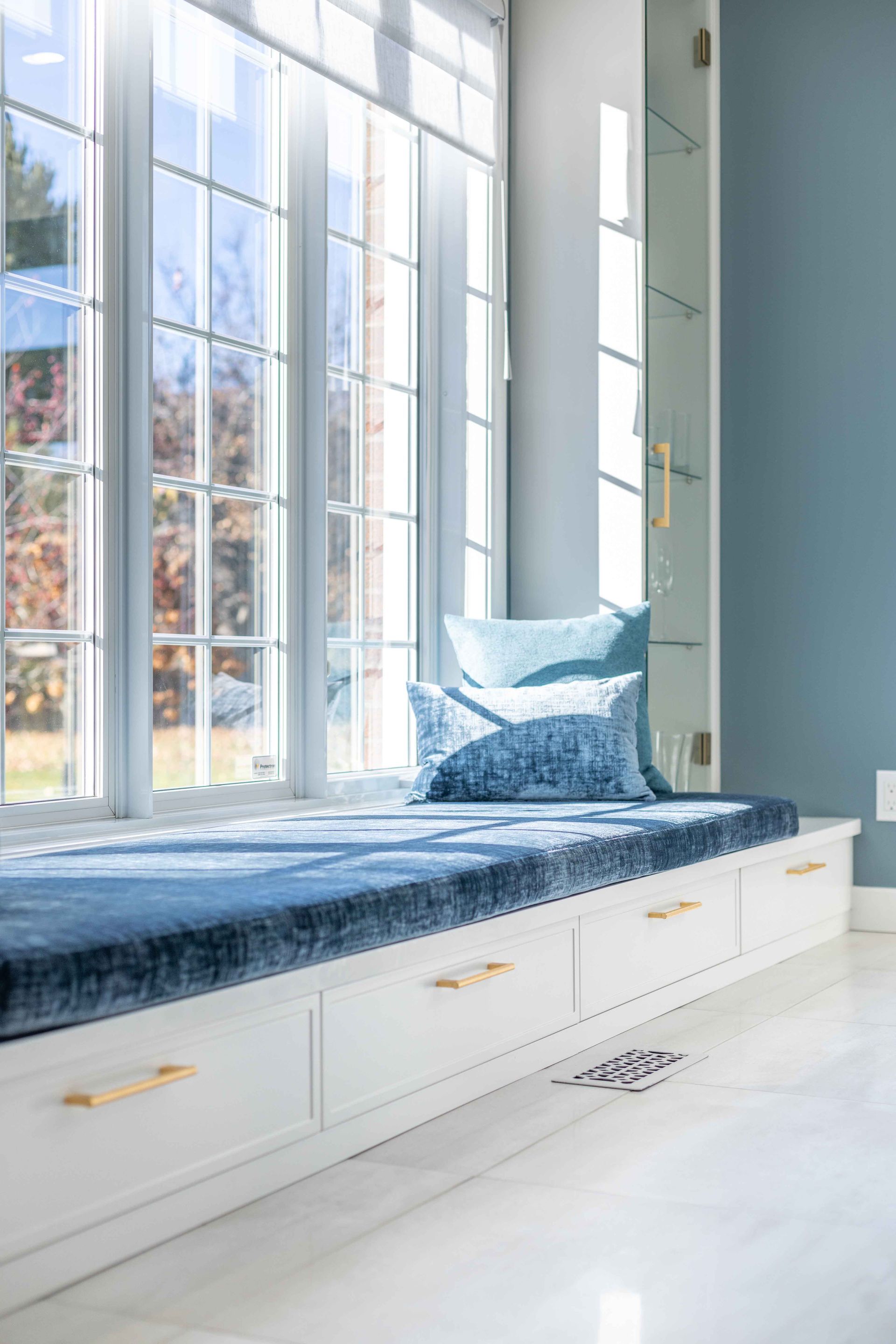 Banquette près de la fenêtre avec coussins bleus et tiroirs blancs, contre une fenêtre ensoleillée, mur turquoise.