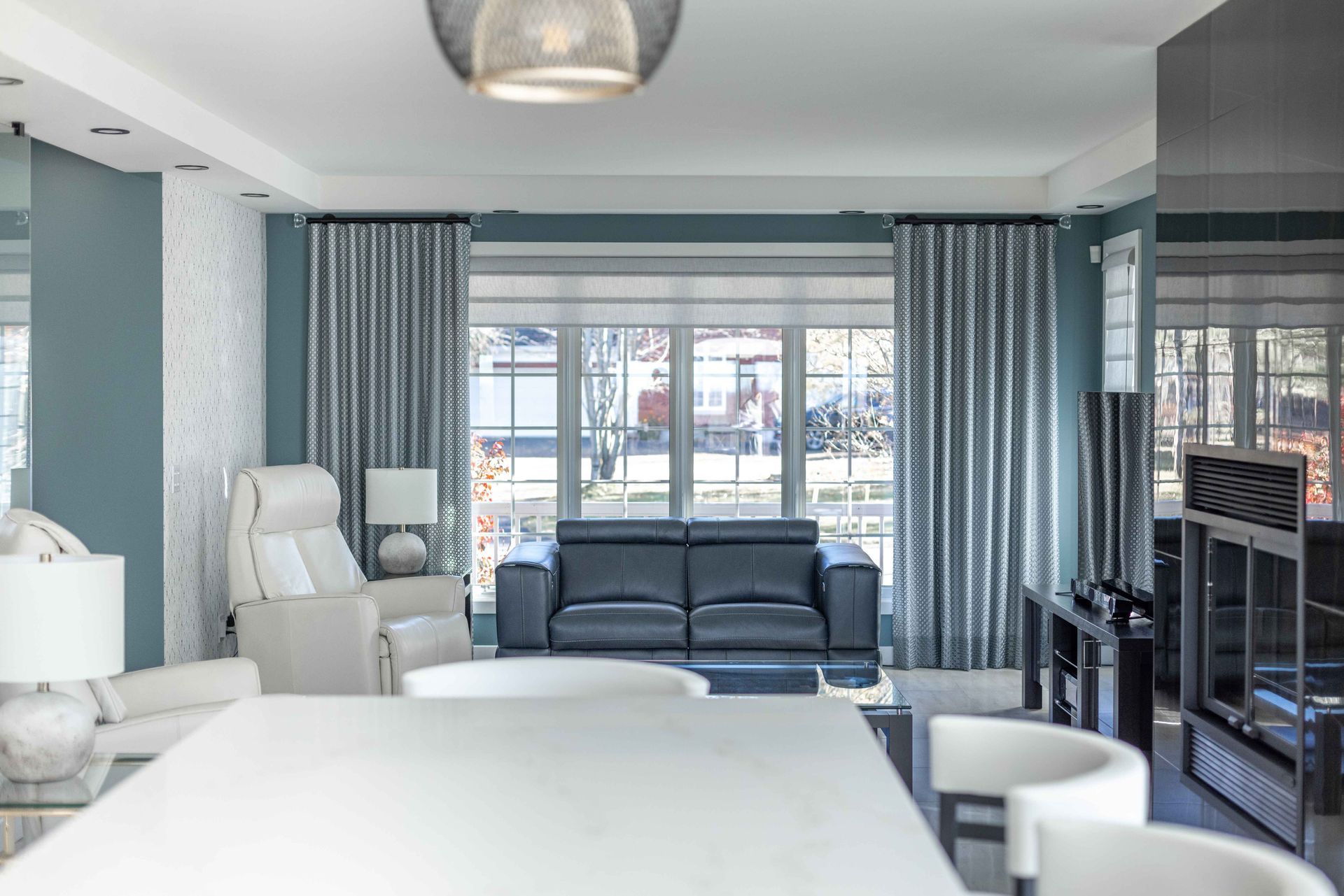 Salon avec murs bleus, rideaux gris et canapé noir devant une fenêtre.