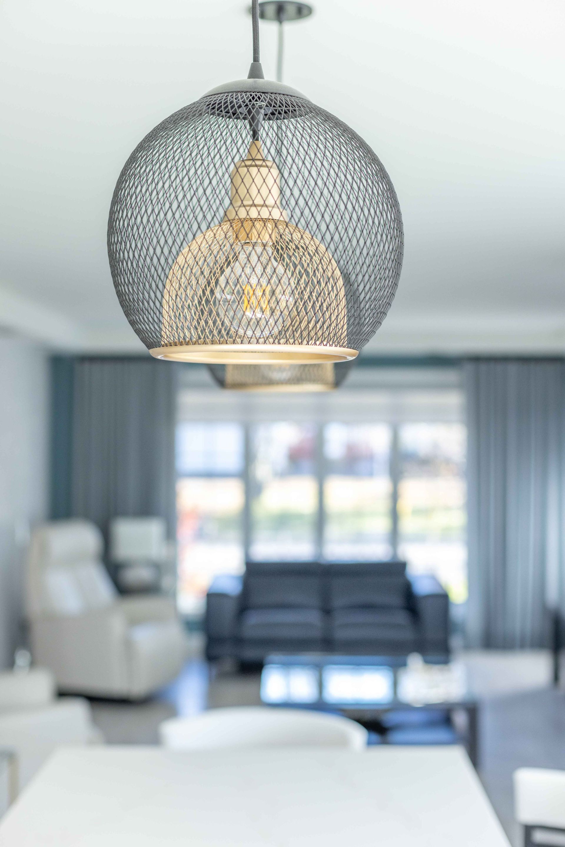 Une suspension lumineuse avec un abat-jour en treillis métallique est installée dans un salon avec un canapé et des fenêtres.
