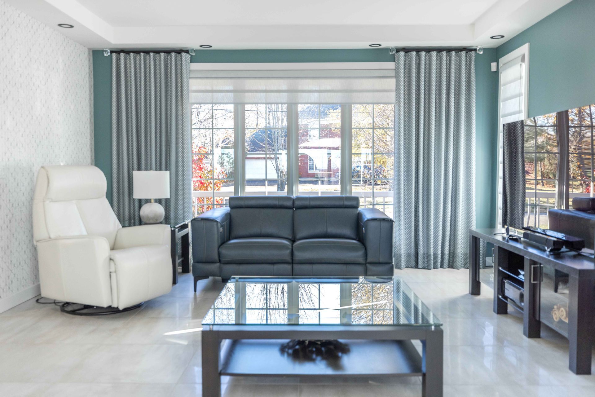 Salon avec rideaux bleu-gris, fauteuil pivotant blanc, canapé noir, table basse en verre et télévision sur un meuble noir.