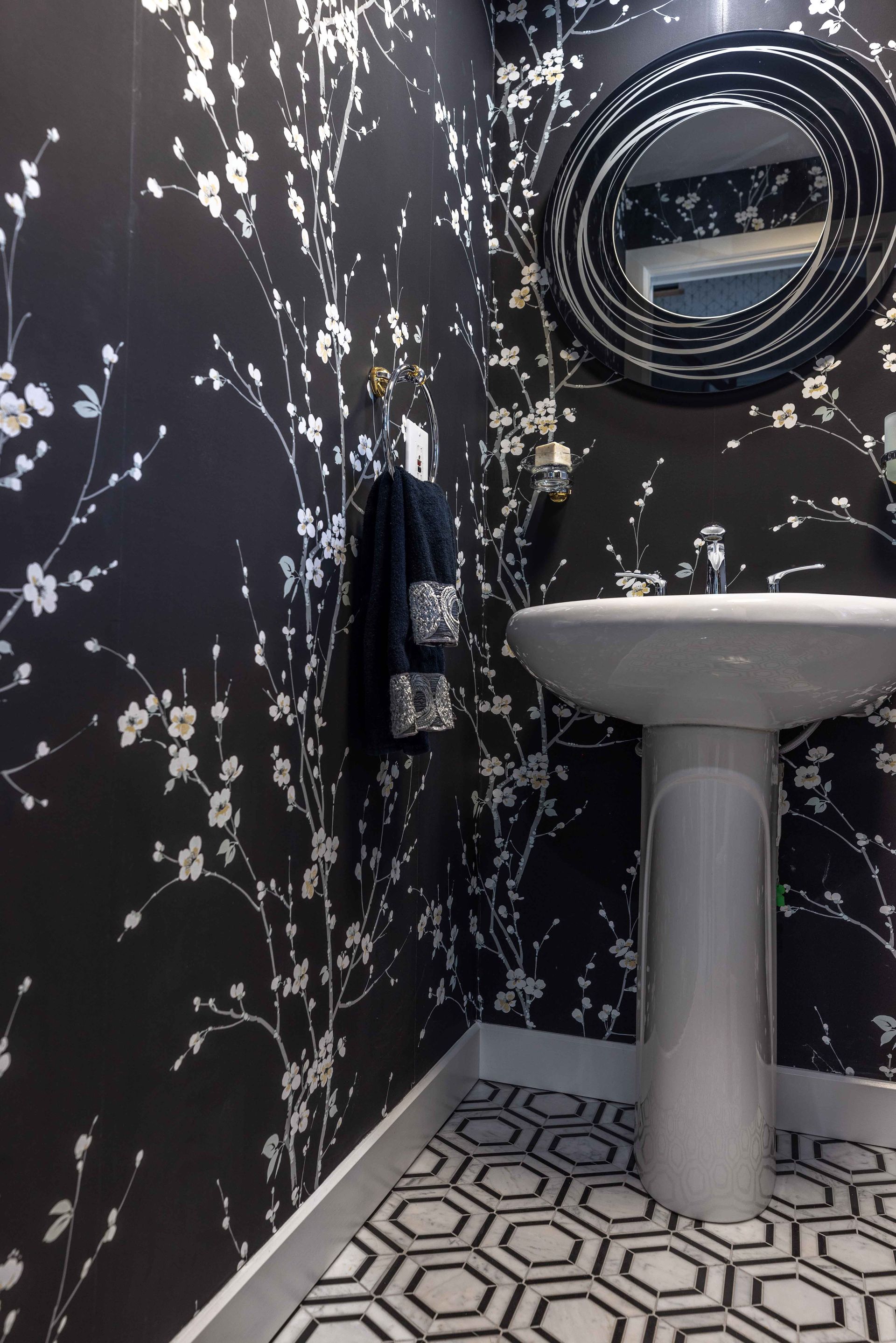Salle de bains avec papier peint floral noir, lavabo sur pied, miroir rond et sol carrelé à motifs.