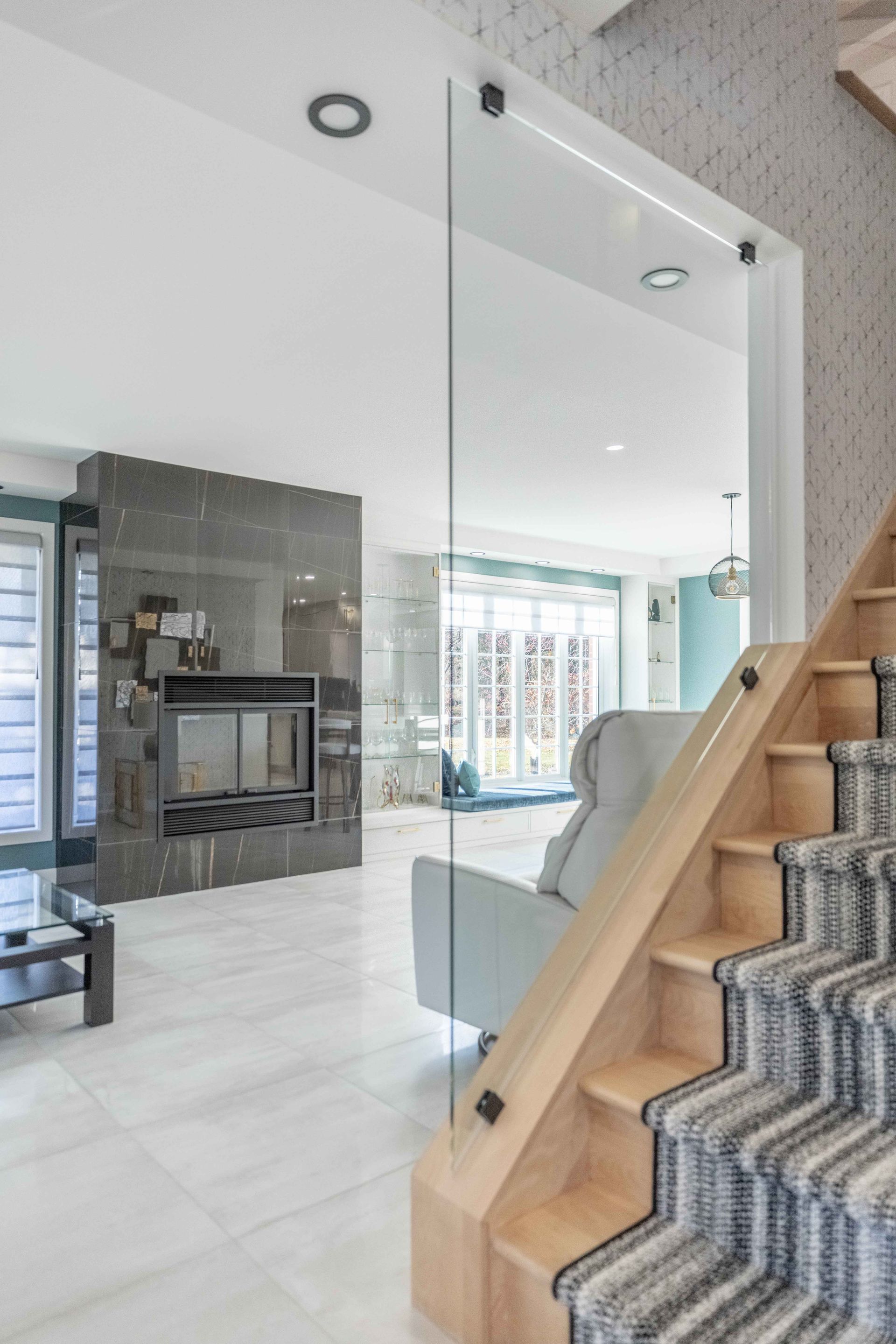 Vue intérieure : salon moderne avec cheminée et escalier, cloison vitrée, tons gris, blancs et bois.