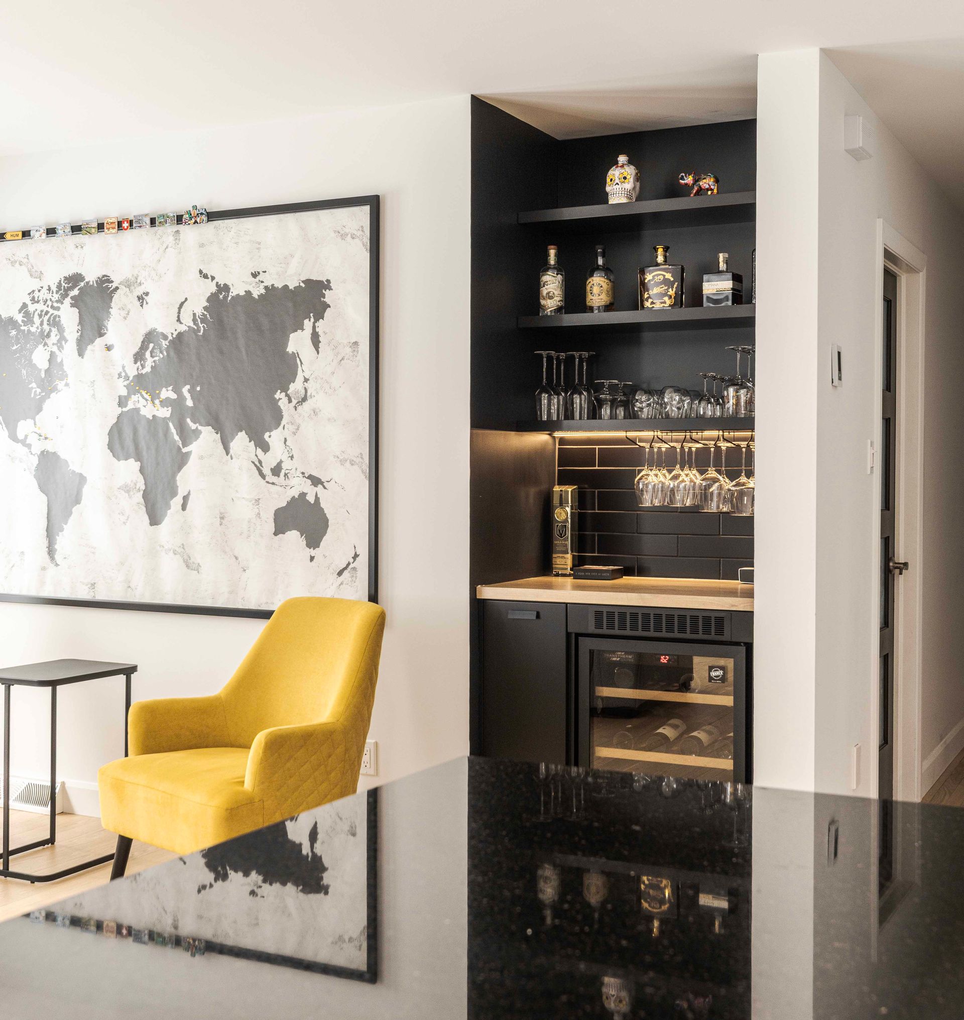 Fauteuil jaune, coin bar avec étagères noires, carte du monde sur un mur blanc et sol noir brillant.