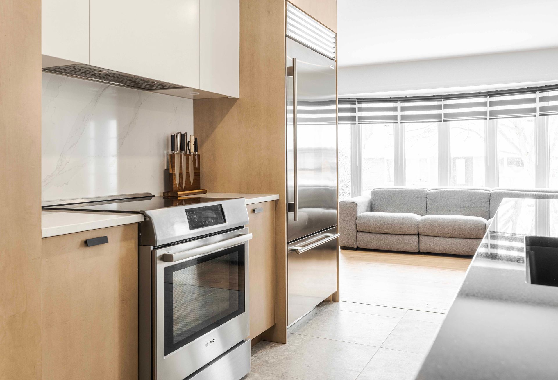 Cuisine moderne avec appareils électroménagers en acier inoxydable, placards en bois clair et coin salon avec canapé gris.