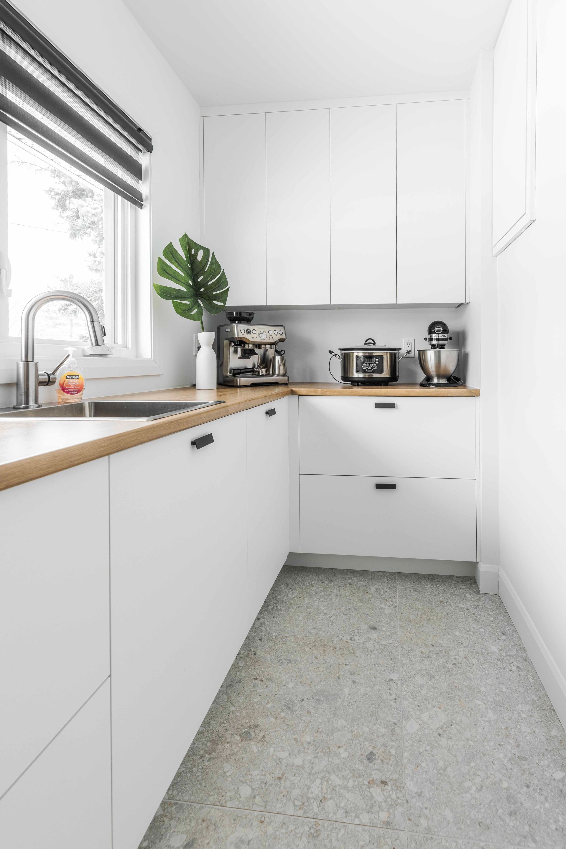 Petite cuisine blanche avec comptoirs en bois, appareils électroménagers et une fenêtre avec un store rayé.
