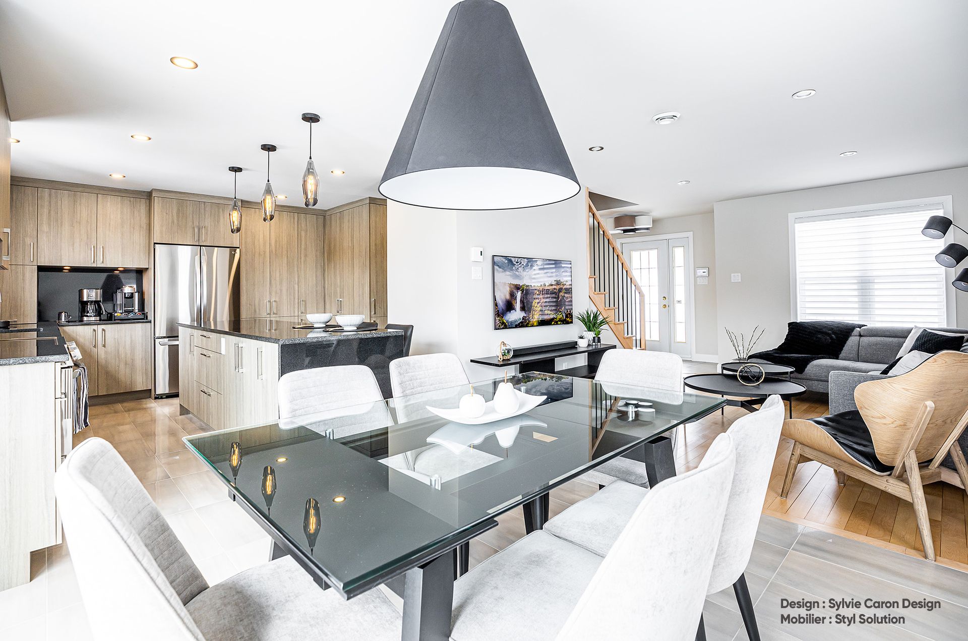 Cuisine et salle à manger à aire ouverte avec table en verre, armoires en bois et suspensions lumineuses grises.