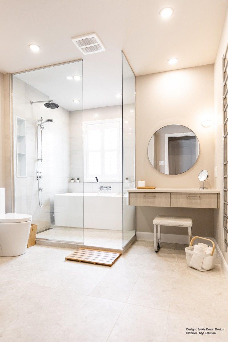 Salle de bains moderne avec douche à l'italienne, baignoire, meuble-lavabo et miroir rond. Murs beiges et robinetterie blanche.