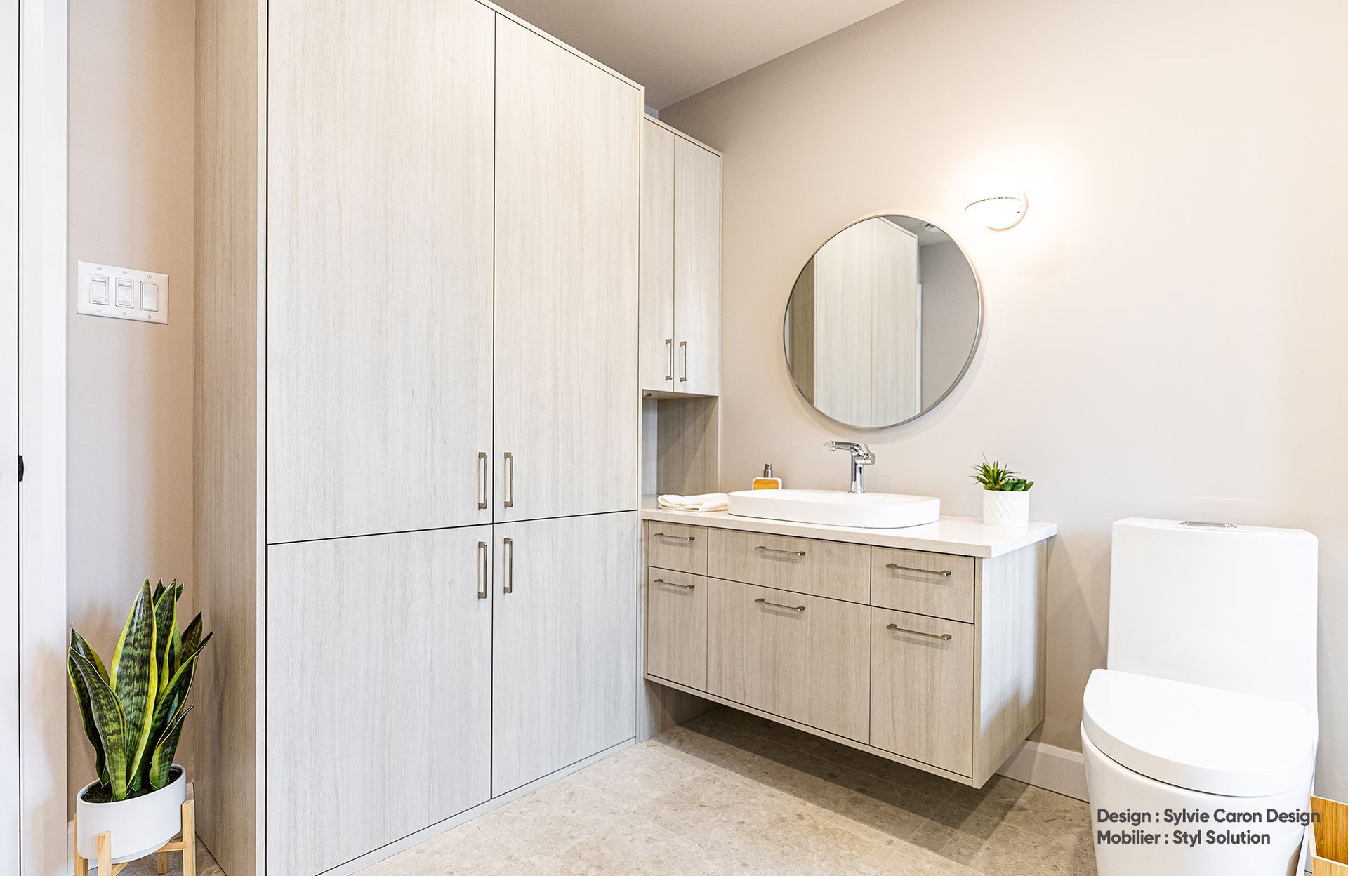 Salle de bains moderne avec meubles en bois clair, miroir rond et toilettes.