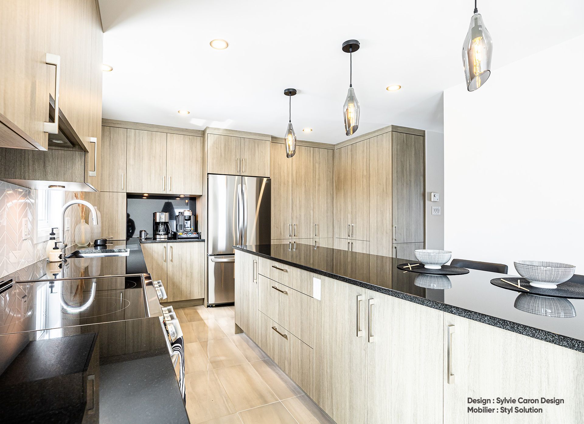 Cuisine avec armoires en bois clair, comptoirs foncés, appareils électroménagers en acier inoxydable et luminaires suspendus.