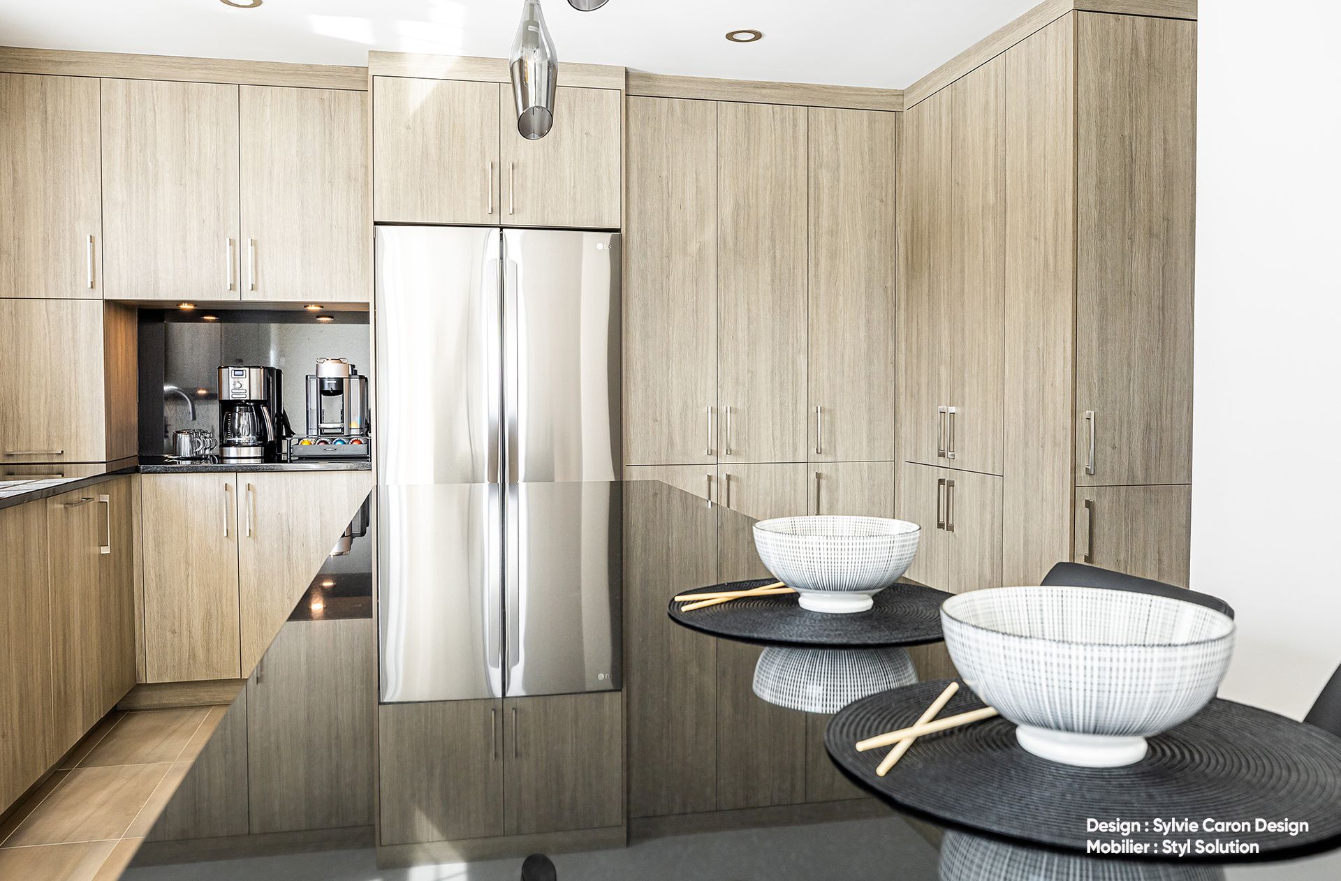 Cuisine moderne avec placards en bois clair, réfrigérateur en acier inoxydable et plan de travail foncé avec bols.
