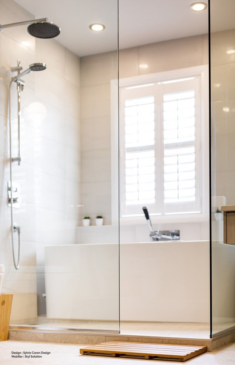 Salle de bains moderne avec douche en verre, murs blancs, fenêtre à volets, tapis de sol en bois.