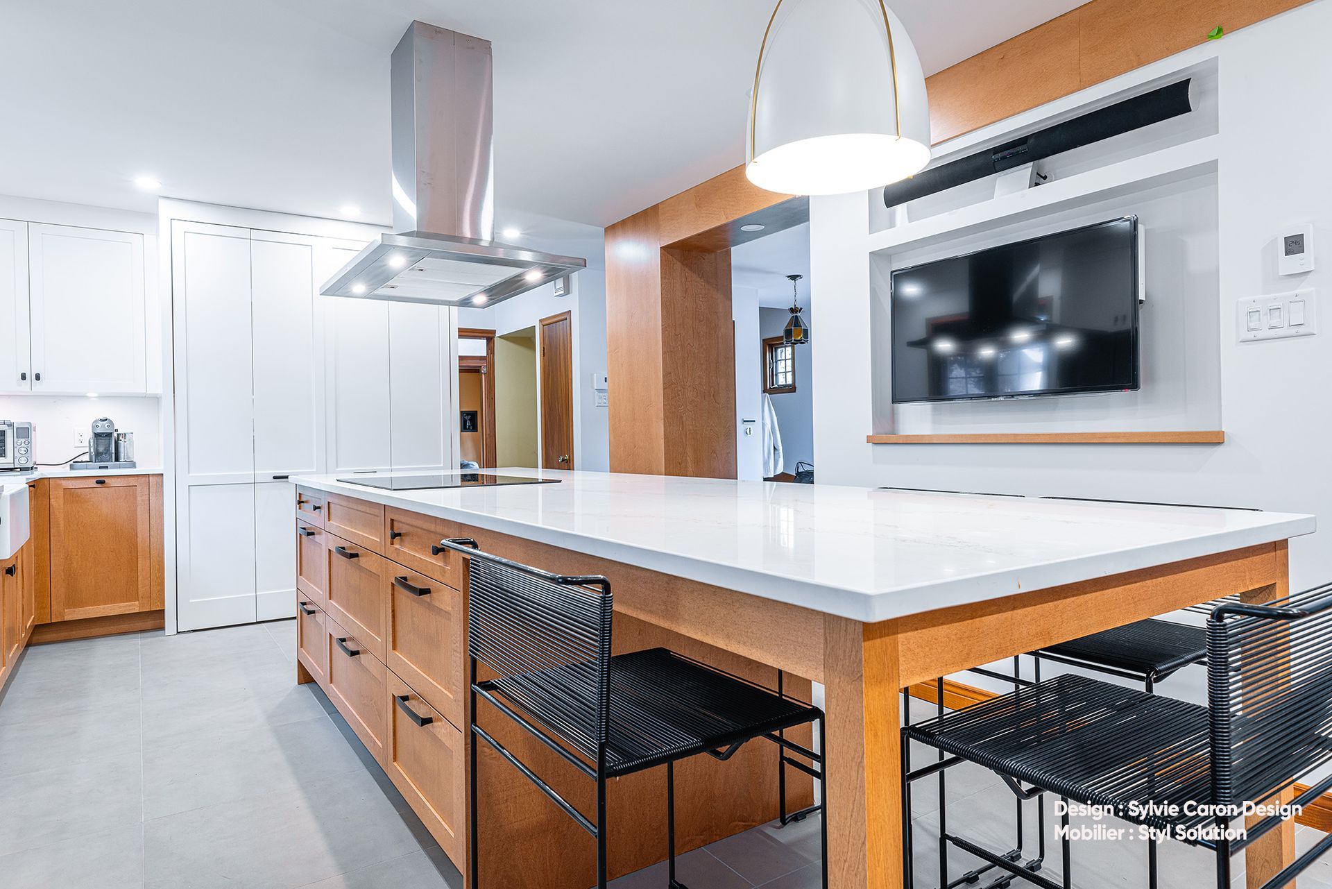 Cuisine moderne avec comptoirs blancs et armoires en bois ; îlot avec chaises noires, hotte aspirante et télévision murale.