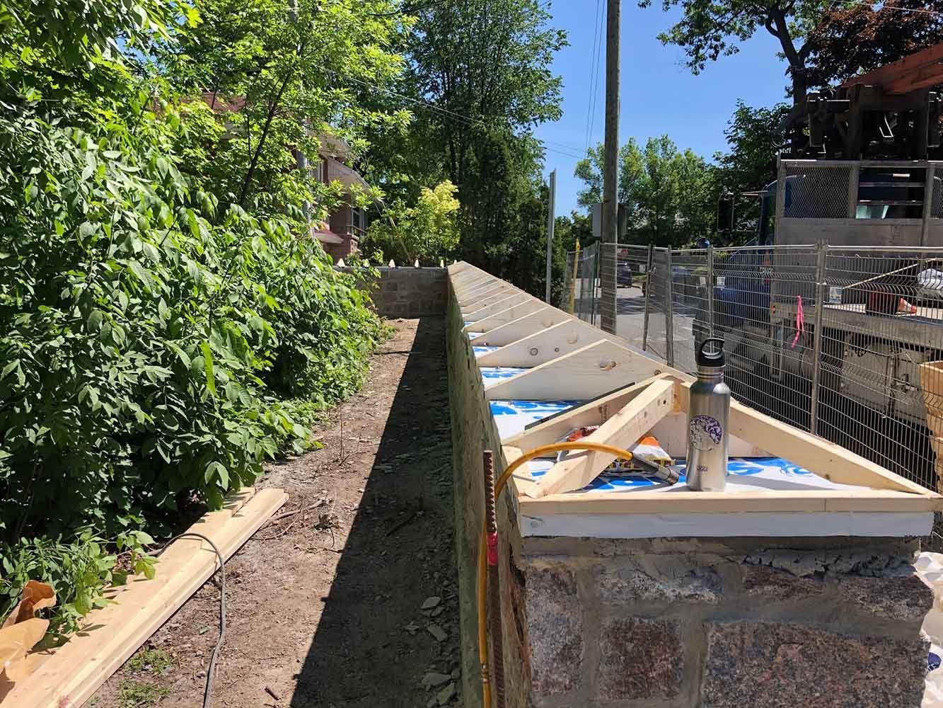 Un mur de soutènement en construction, avec des supports en bois, à côté de verdure par une journée ensoleillée.