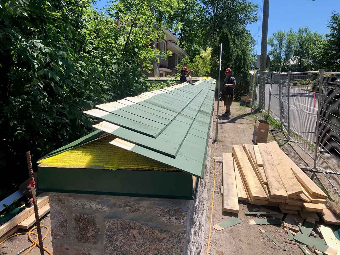 Des ouvriers installent des tuiles sur une structure en pierre ; on aperçoit les matériaux de construction.