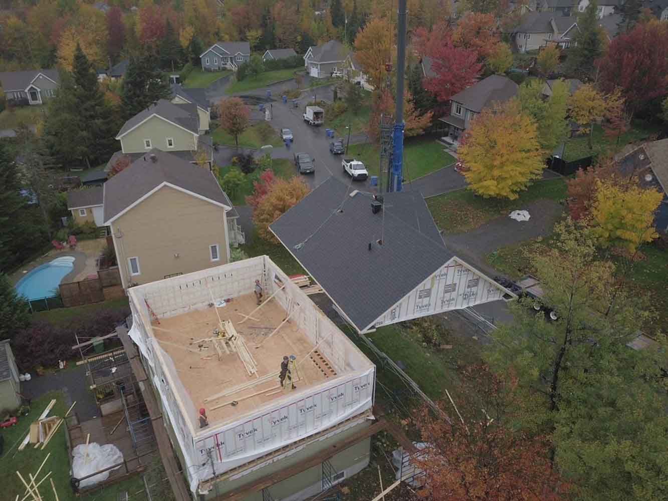Vue aérienne : une maison en construction avec une grue soulevant une partie du toit ; arbres d’automne et maisons de banlieue.