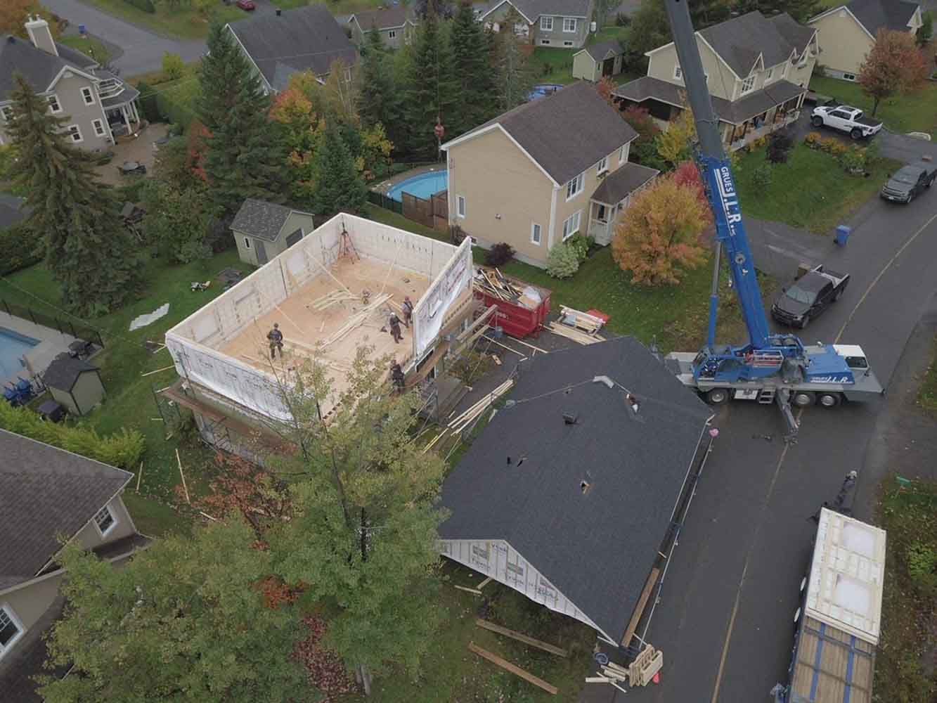 Une grue soulève une partie d'une maison en construction au-dessus d'un petit bâtiment dans une rue résidentielle.
