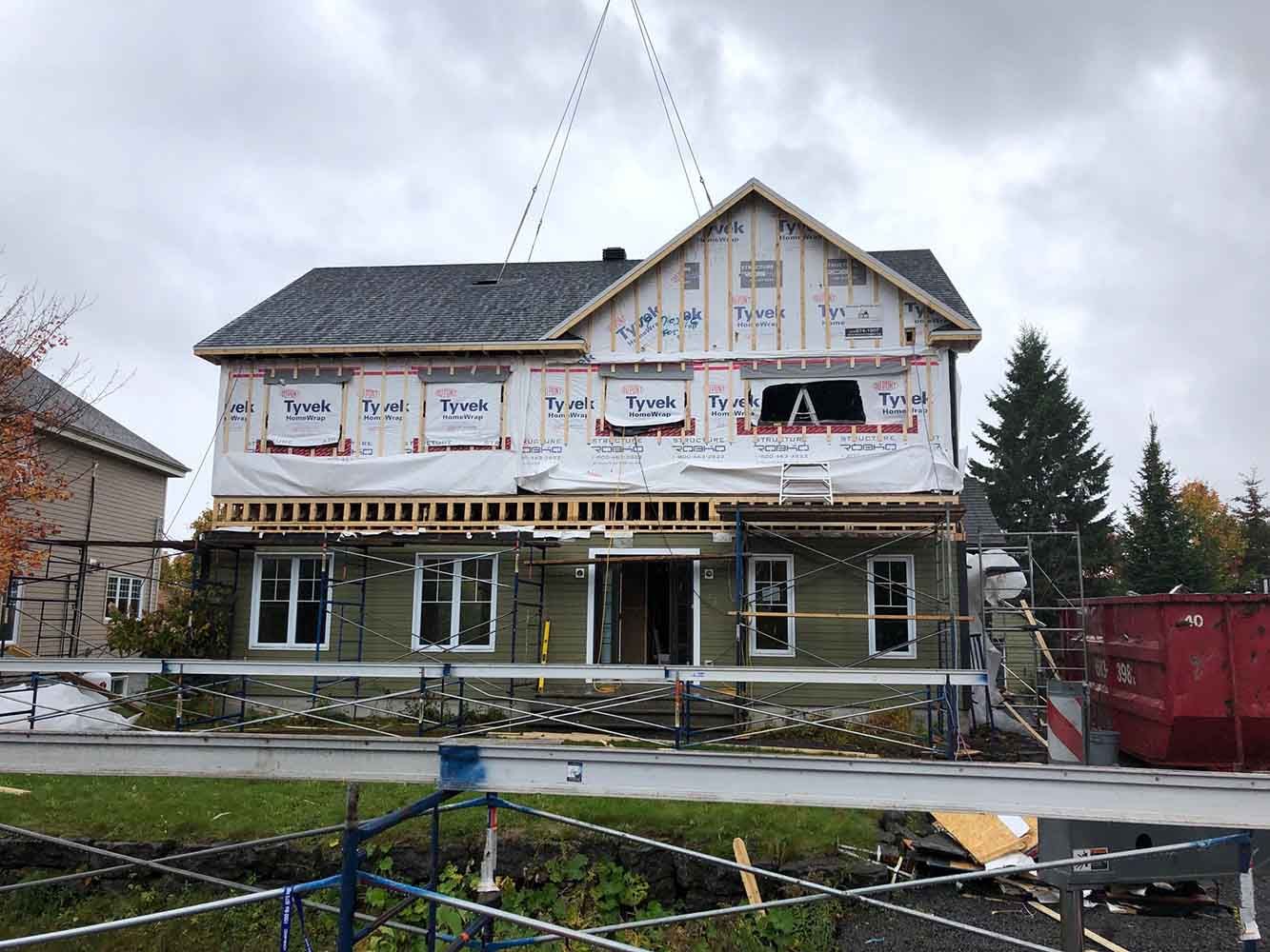 Maison à deux étages en construction, recouverte de bâches Tyvek et d'échafaudages, soulevée par une grue.
