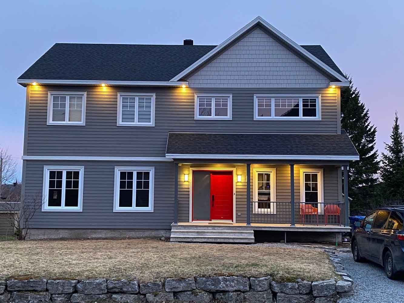 Maison grise à deux étages avec une porte rouge, un porche éclairé et un jardin aménagé au crépuscule.