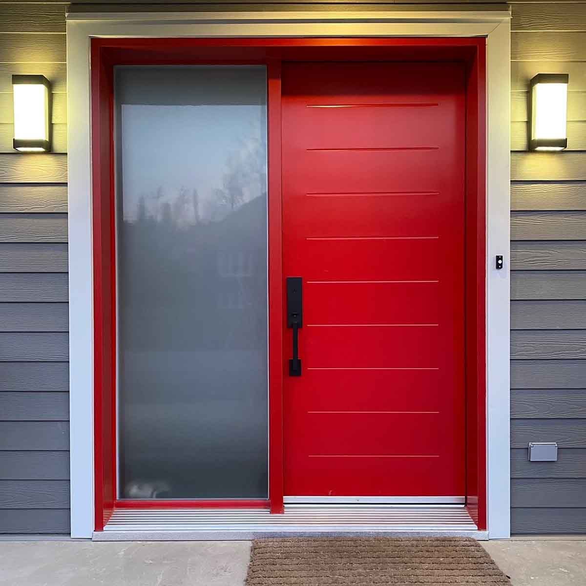 Porte d'entrée rouge avec panneau en verre dépoli, flanquée d'appliques murales sur une façade grise.