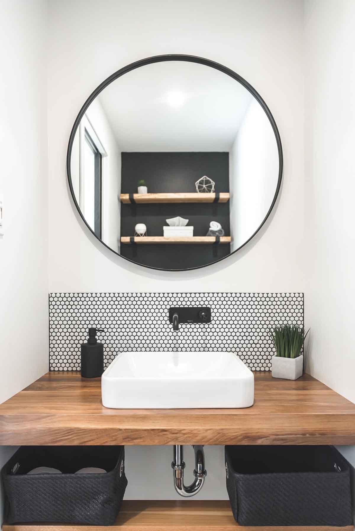 Salle d'eau avec comptoir en bois, lavabo carré, miroir rond et crédence à motifs.