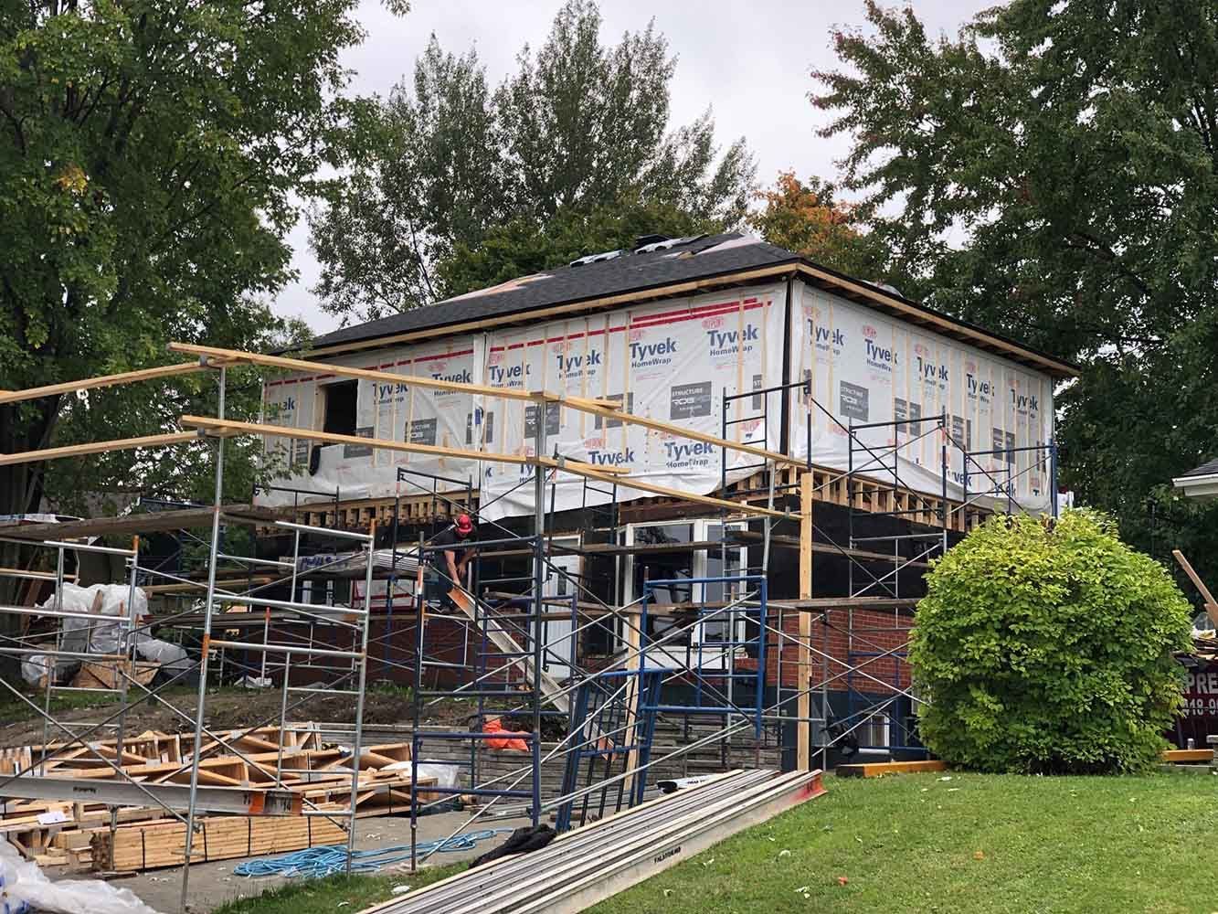 Maison en construction avec échafaudages. Murs extérieurs recouverts de Tyvek. Arbres et pelouse en arrière-plan.