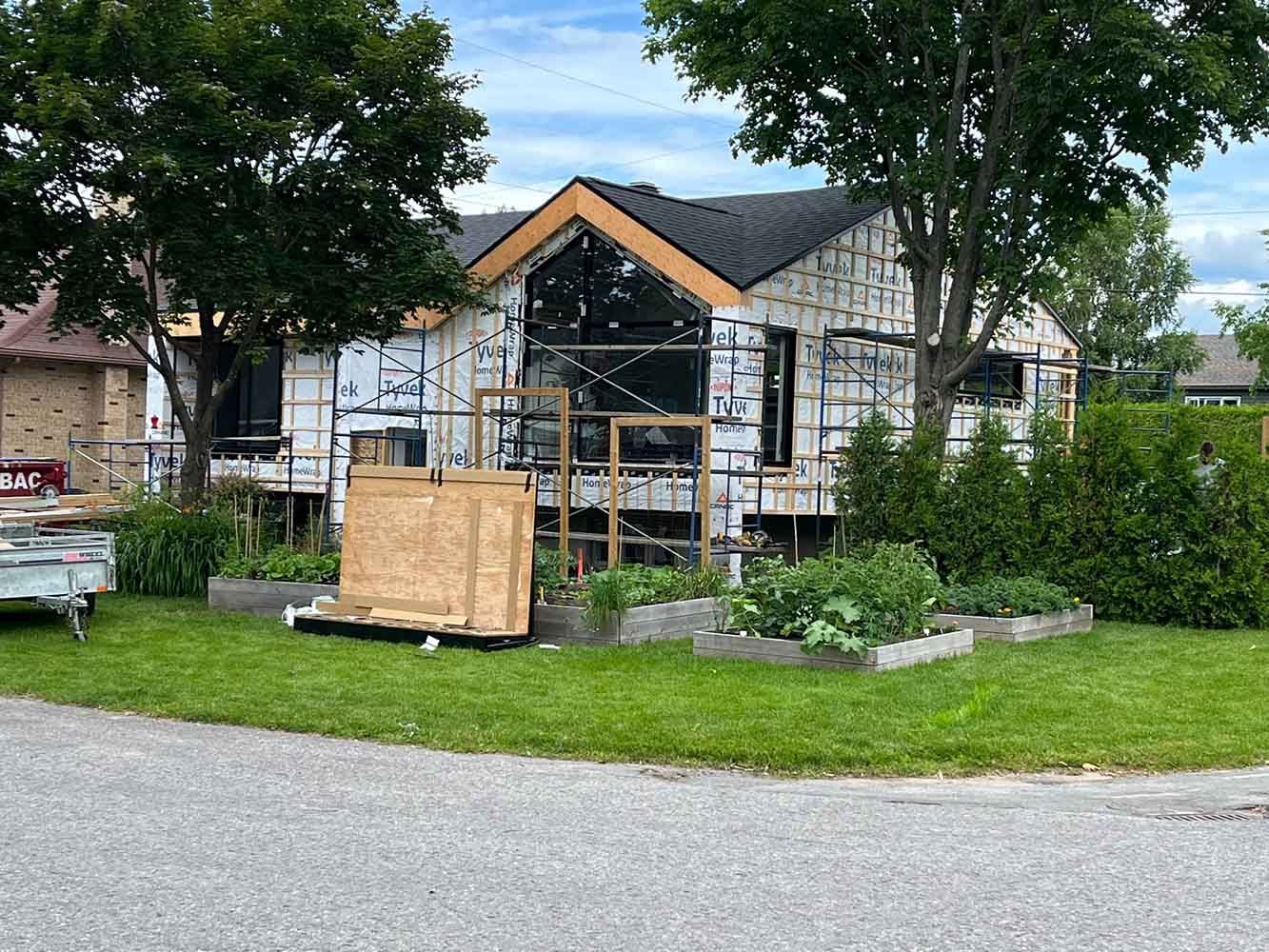 Maison en construction, vue extérieure. Bardage blanc, toit noir, ossature bois, jardin devant.