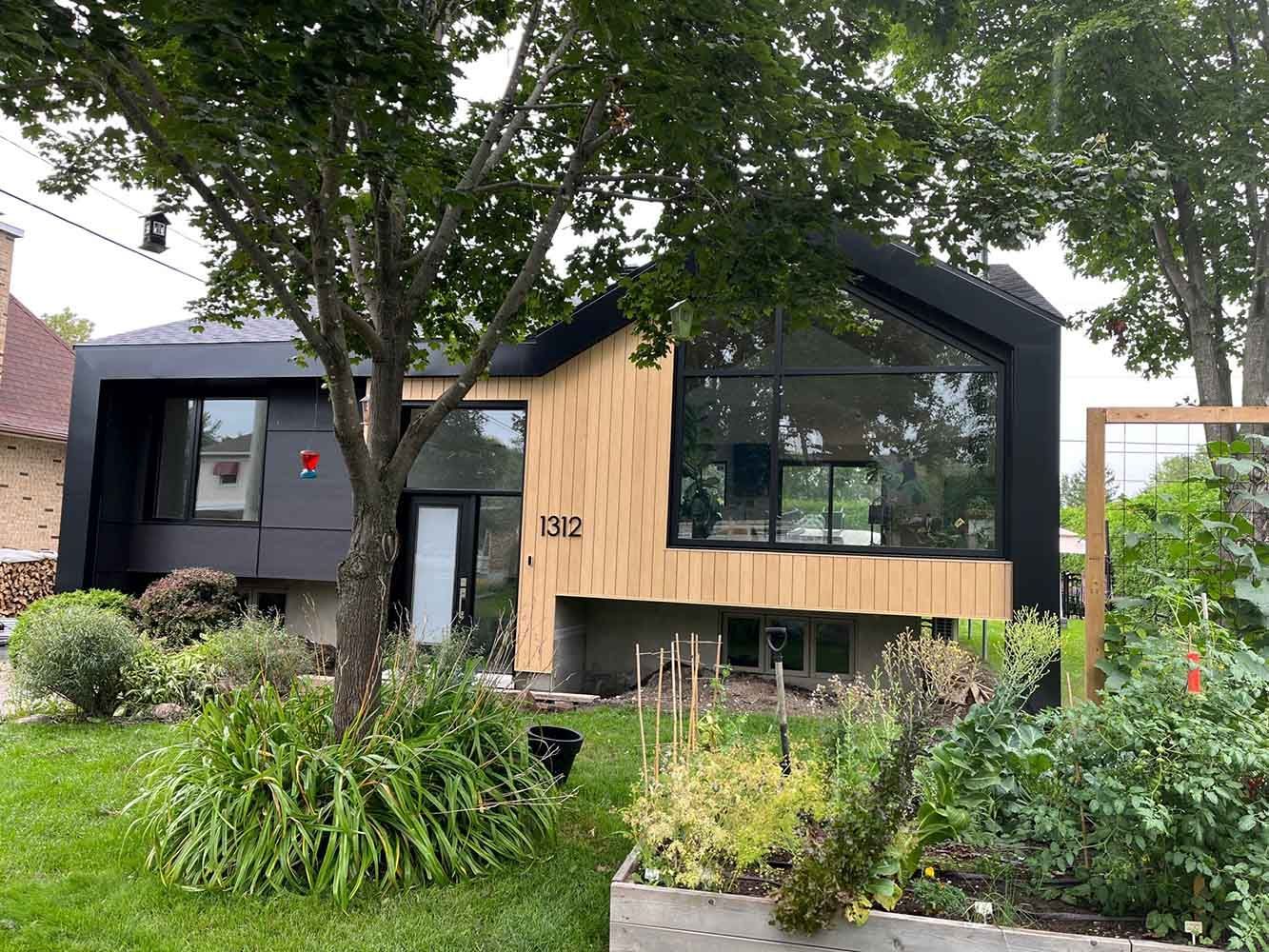 Maison moderne avec bardage noir et beige, grandes fenêtres et jardin à l'avant.