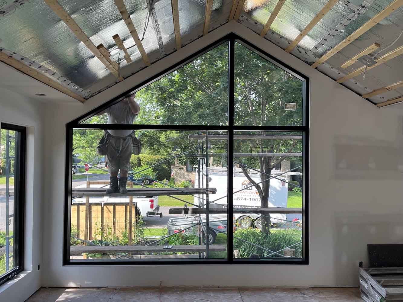 Vue intérieure d'un bâtiment avec une grande fenêtre. Une personne se trouve sur un échafaudage à l'extérieur.