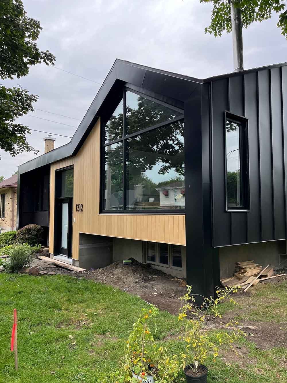 Extérieur de maison moderne avec bardage noir et bois, grande fenêtre et pelouse verte.
