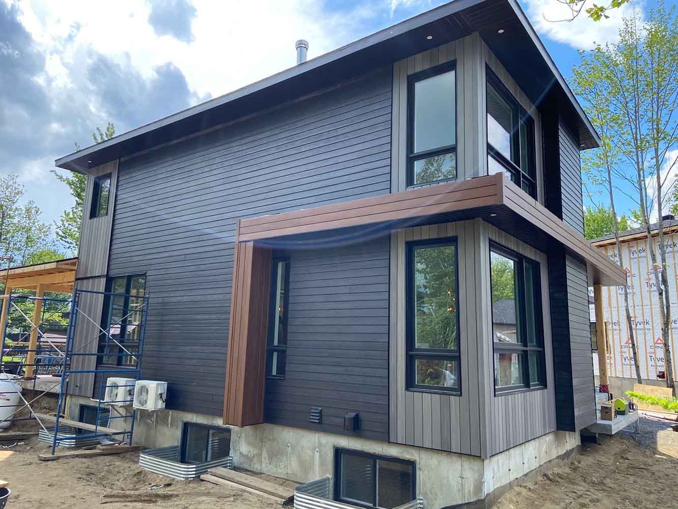 Maison moderne à deux étages avec bardage gris, fenêtres à cadre noir et boiseries. Matériaux de construction apparents.