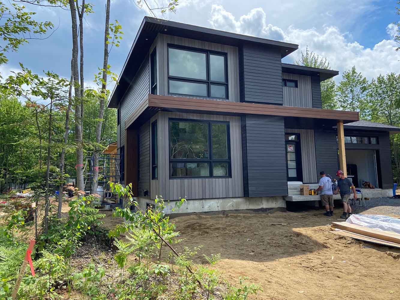 Maison moderne à deux étages avec un revêtement gris. Des hommes travaillent sur le porche sous un ciel nuageux.
