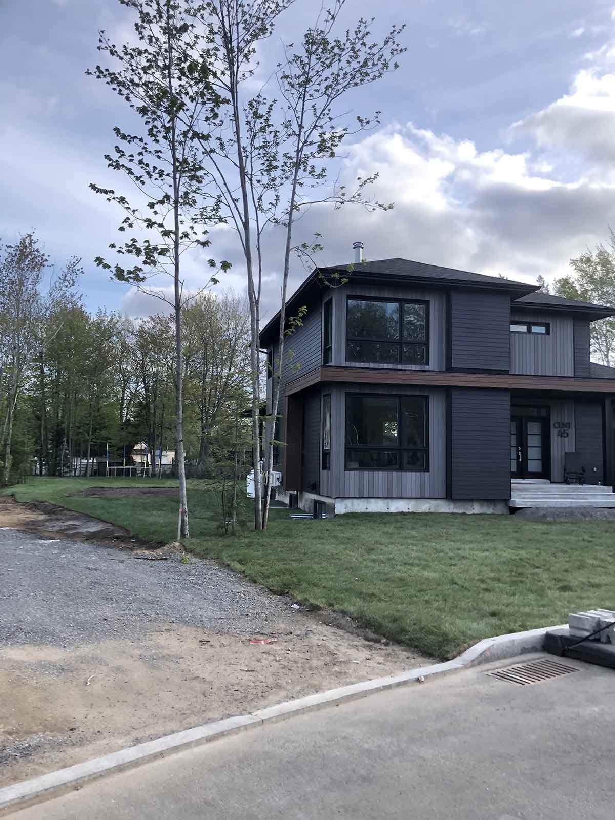 Maison moderne à deux étages avec bardage gris, grandes fenêtres et pelouse. Arbres à l'avant et allée gravillonnée.