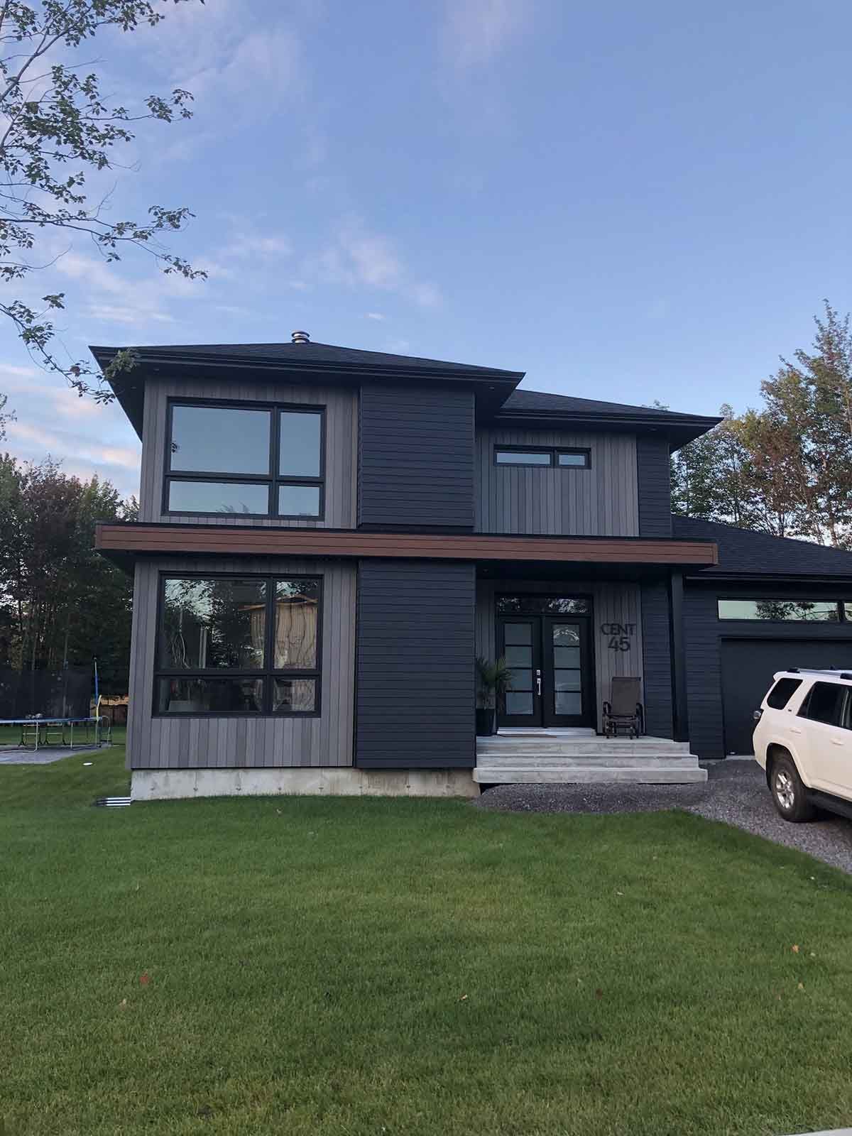 Maison moderne à deux étages avec un revêtement extérieur gris foncé, de grandes fenêtres et un SUV blanc garé dans l'allée.