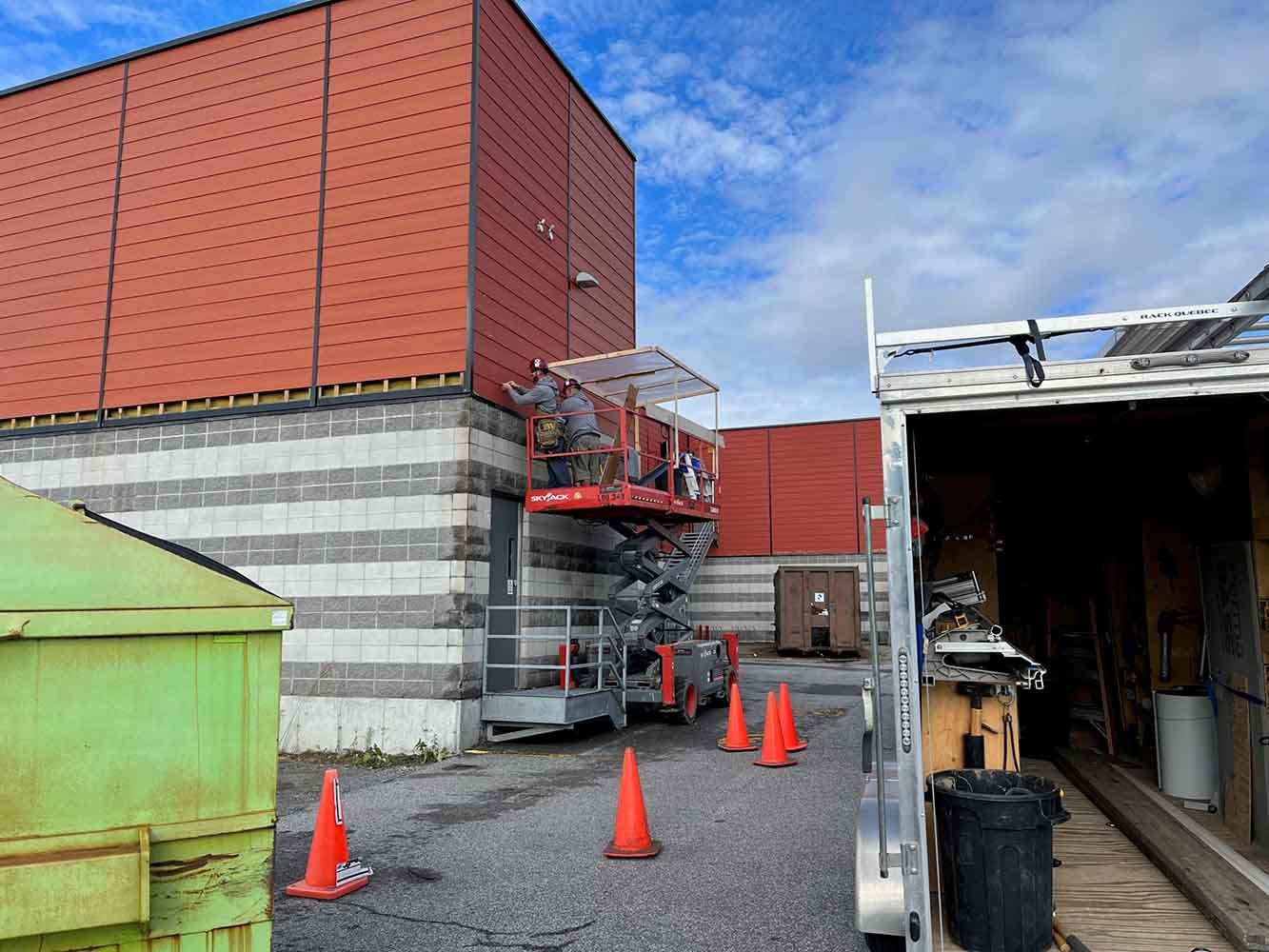 Des ouvriers du bâtiment réparent la façade rouge d'un immeuble depuis une nacelle élévatrice. Des cônes orange et une remorque se trouvent à proximité.